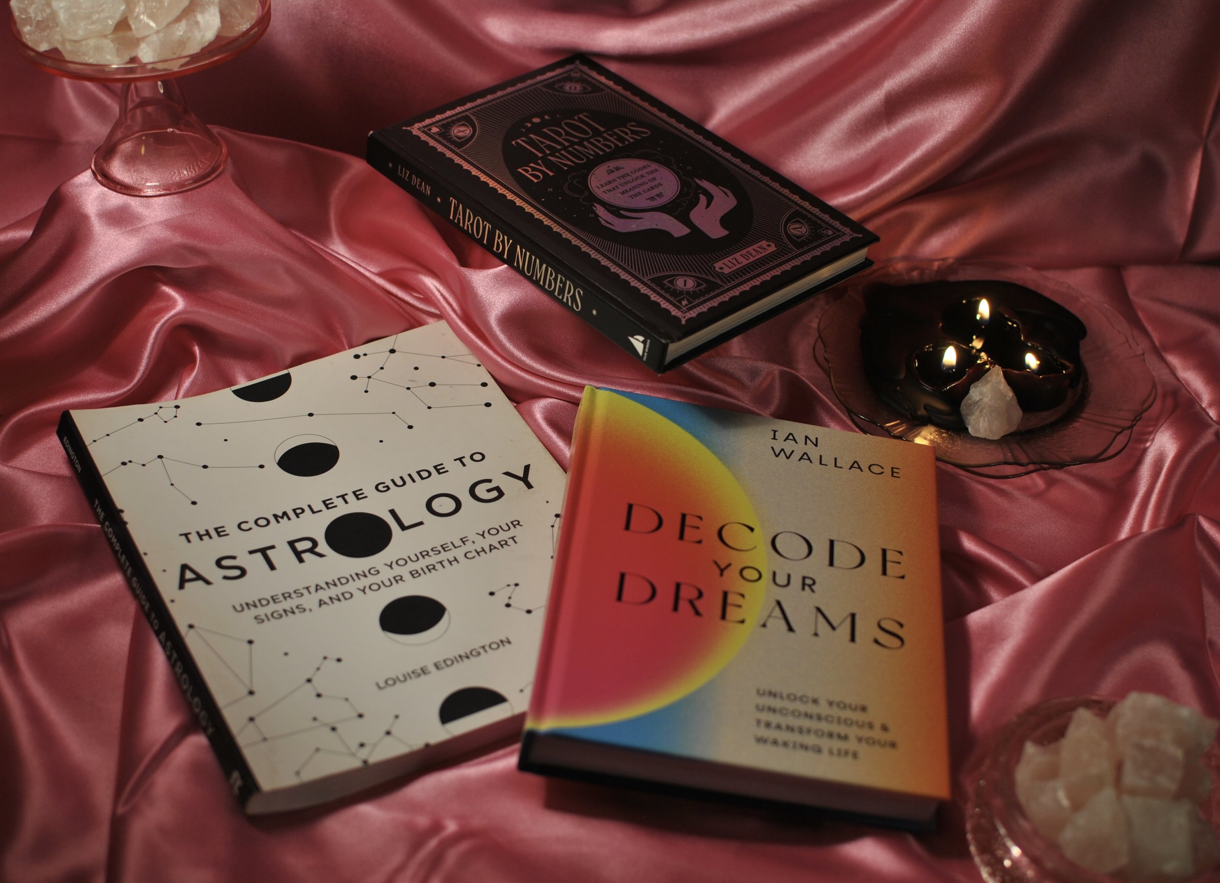 the three books mentioned in the post about tarot, astrology, and dreamwork are photographed here with a pink satin backdrop, rose quartz, and black candles.