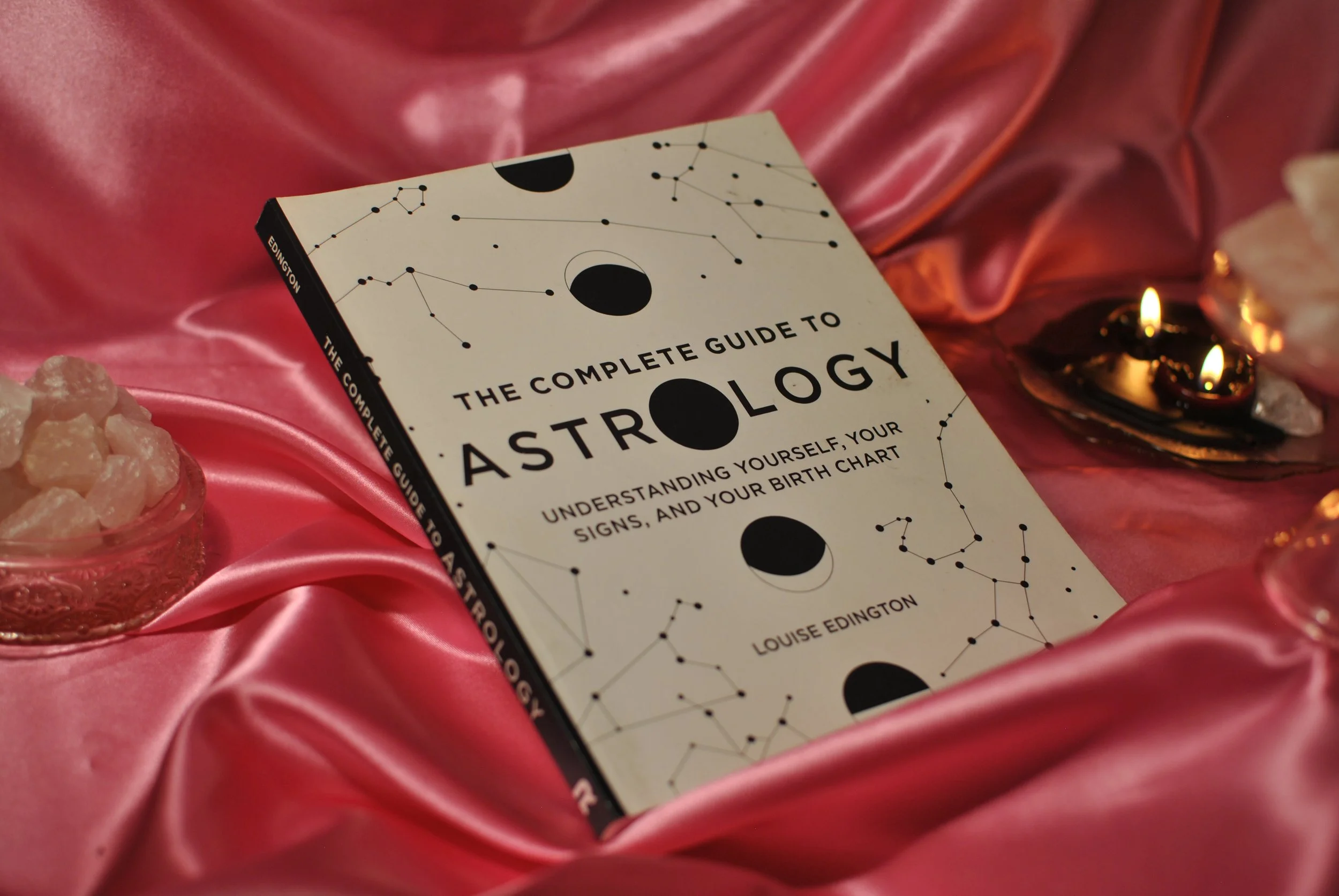 "the complete guide to astrology: understanding yourself, your signs, and your birth chart" by louise edington is photographed here with a pink satin backdrop, rose quartz, and black candles.