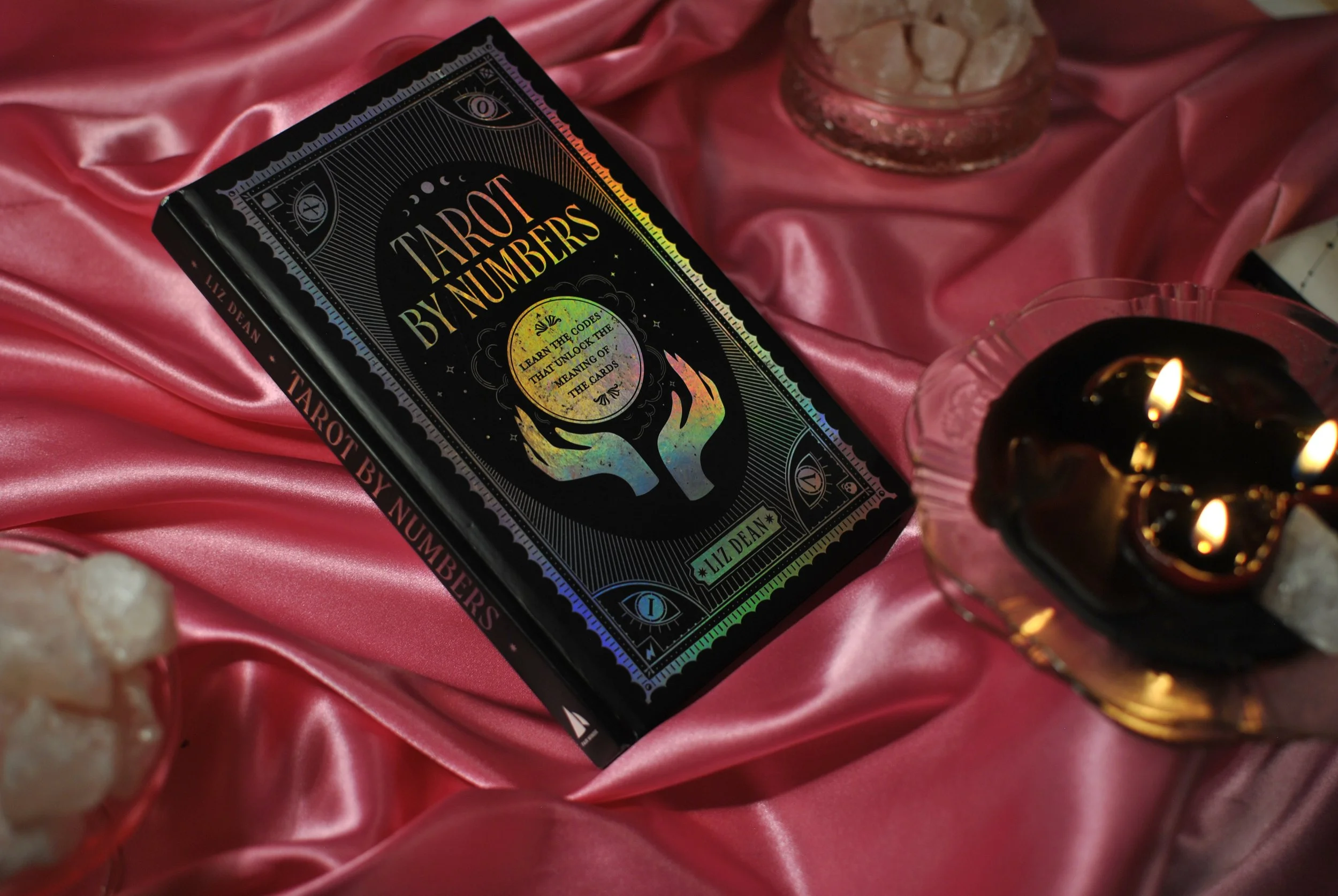 "tarot by numbers," a book by liz dean, is photographed here with a pink satin backdrop, rose quartz, and black candles.