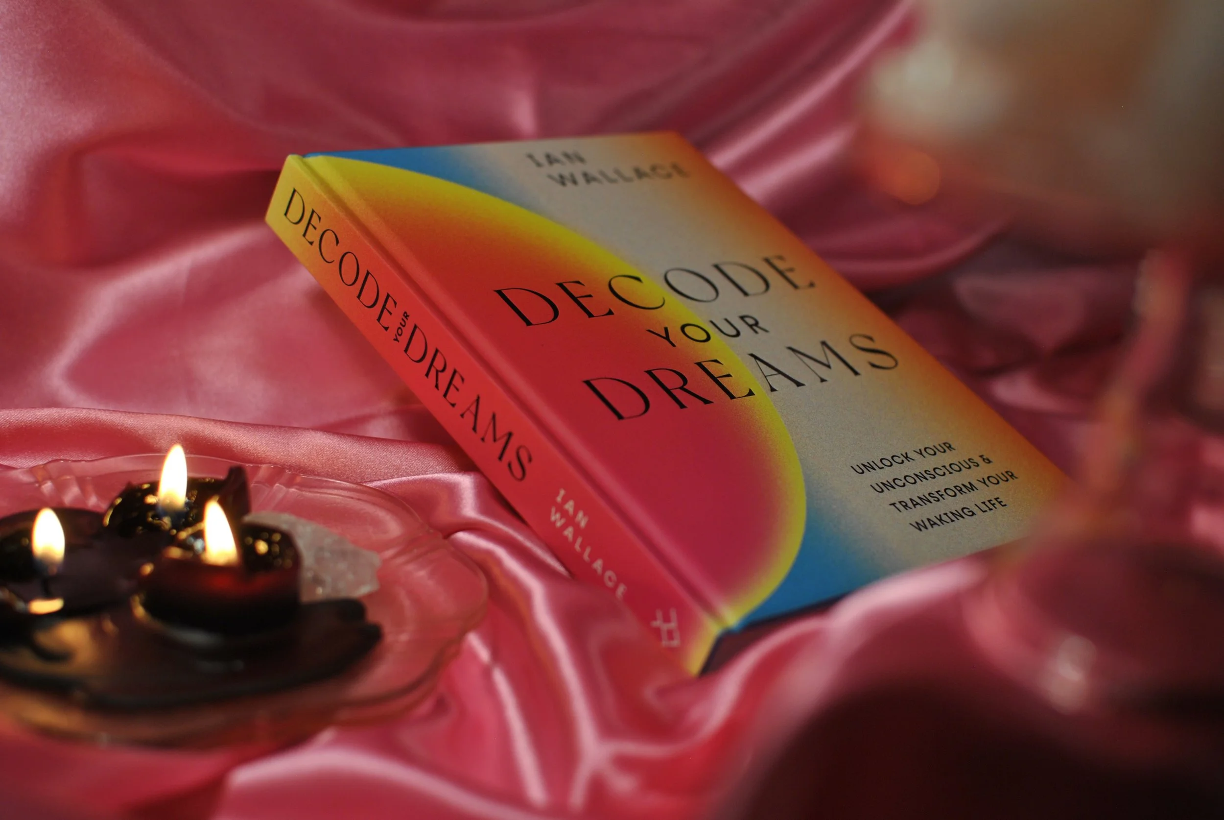 "decode your dreams," a book by ian wallace via white lion publishing, is photographed here with a pink satin backdrop, rose quartz, and black candles.