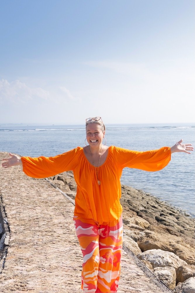 Anastasia Kaesbach trägt ein orangefarbenes Oberteil und eine bunt gemusterte Hose und steht mit ausgestreckten Armen und einem Lächeln auf einem Steinweg am Meer.