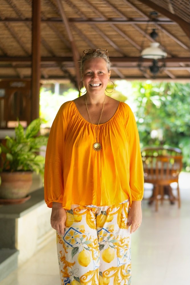 Anastasia Kaesbach in gelbem Oberteil und zitronengelber Hose auf einer Veranda mit Grünpflanzen und Holzmöbeln im Hintergrund.