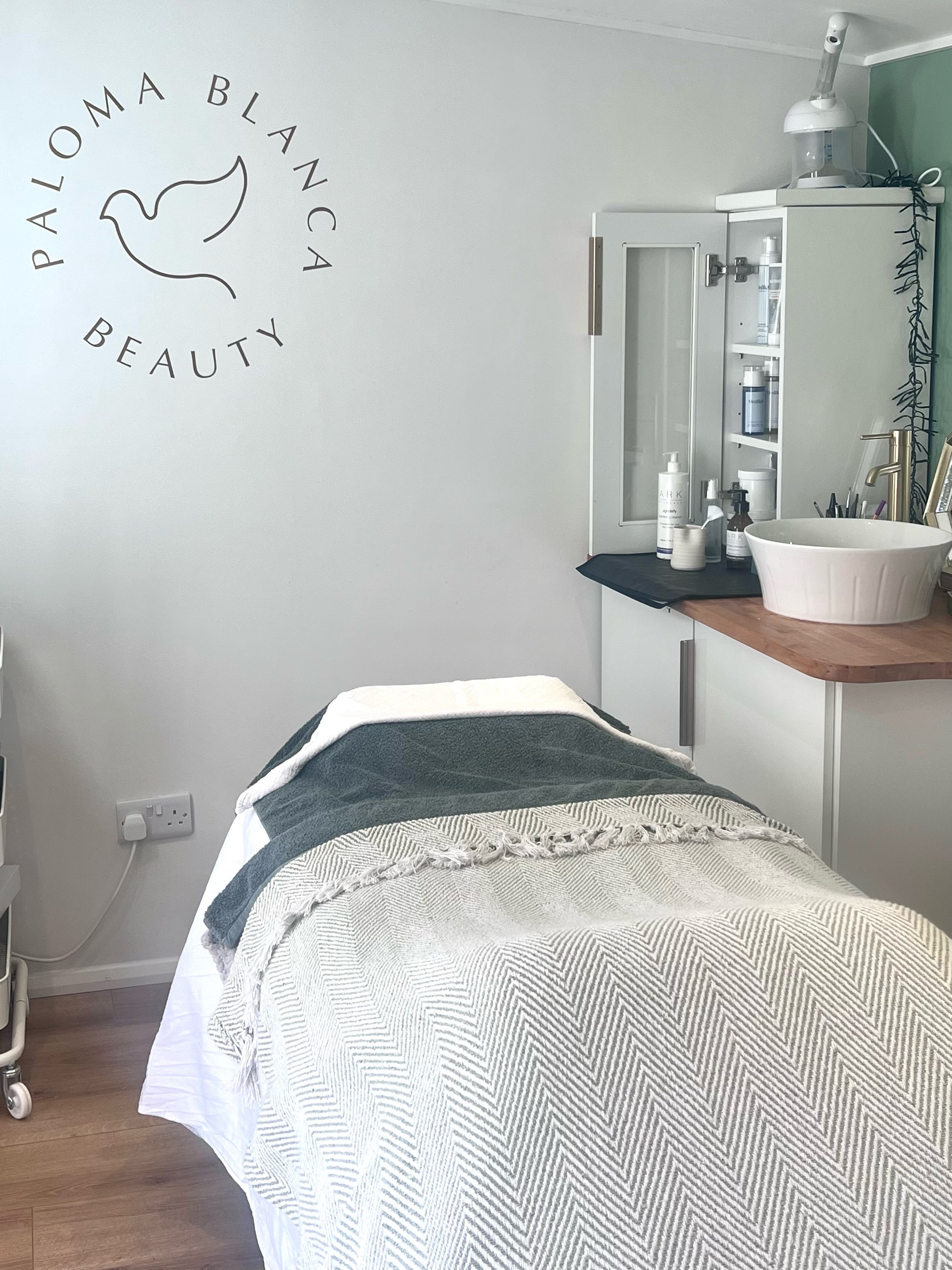 A treatment room at Paloma Blanca Beauty, featuring a massage table covered with a patterned blanket and towels. The room has a cabinet with skincare products and equipment.