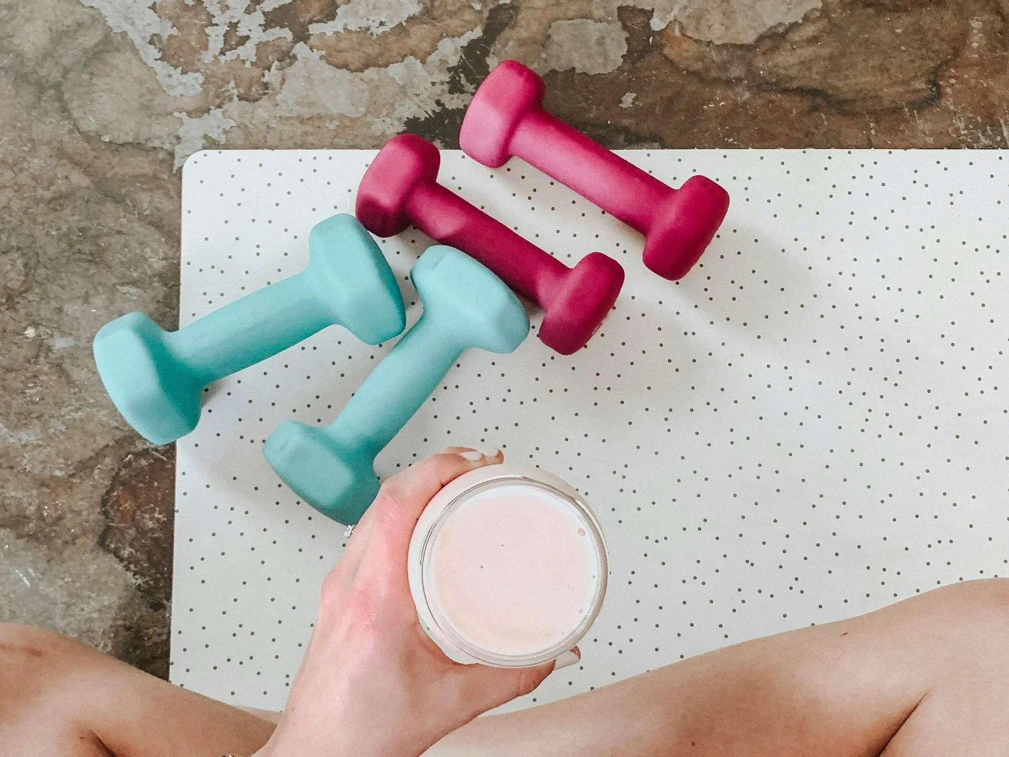 view of a yoga mat and two sets of dumbbells and a human holding a smoothie