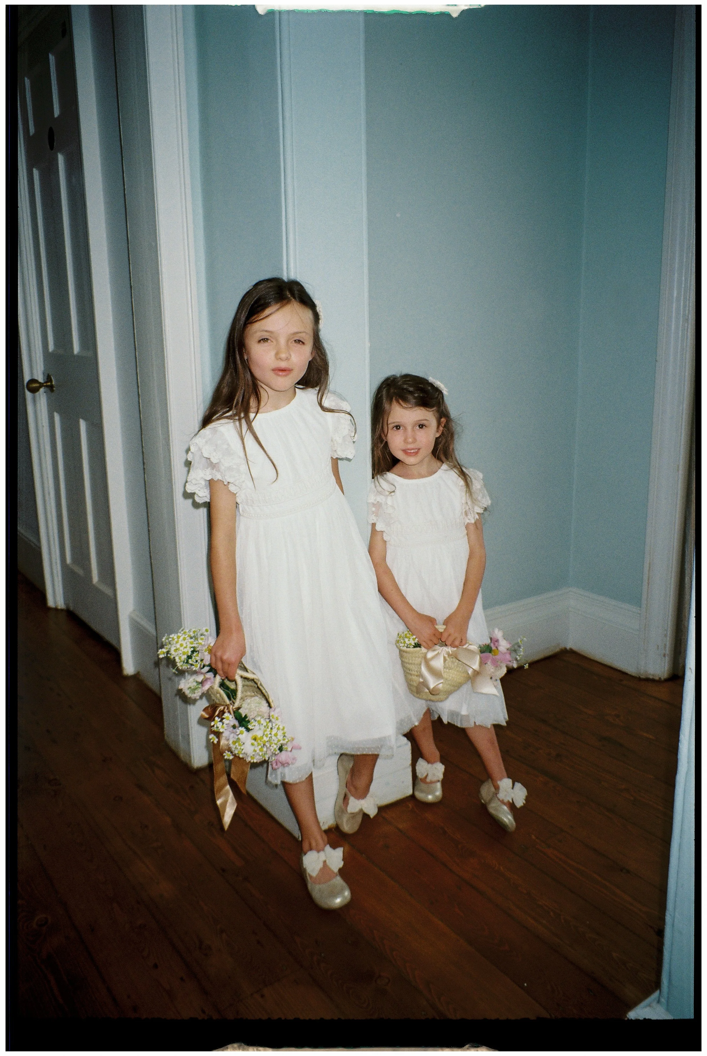 Two young girls in white dresses standing indoors, holding baskets of flowers, likely dressed for a wedding or special occasion.