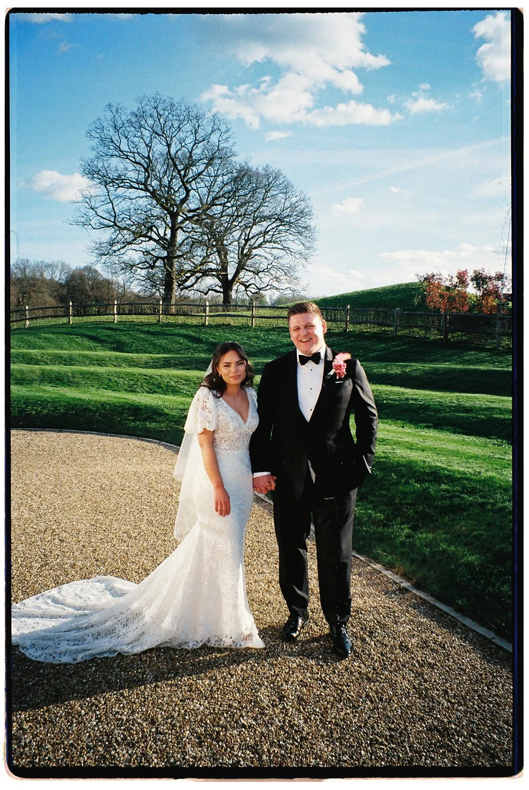 Bride and groom holding hands outdoors on wedding day, with green grass, trees, and blue sky with clouds in the background.