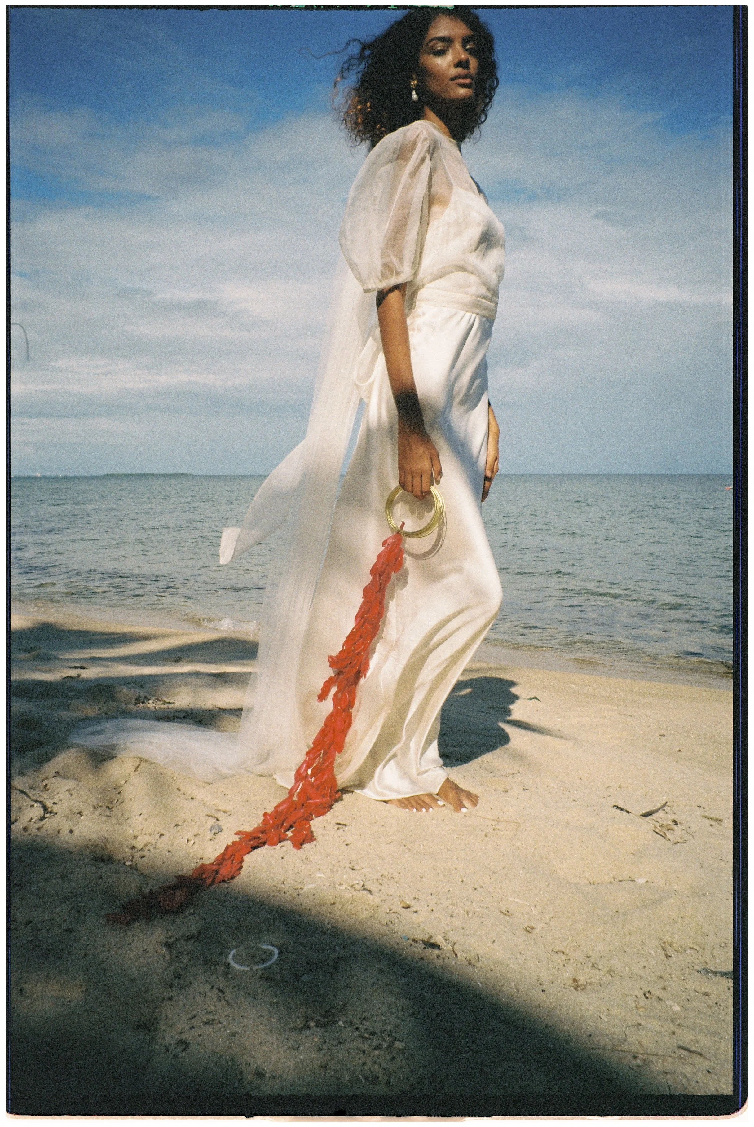 A woman in a flowing white dress standing barefoot on a beach, holding a red rope and gold rings, with the ocean and cloudy sky in the background.