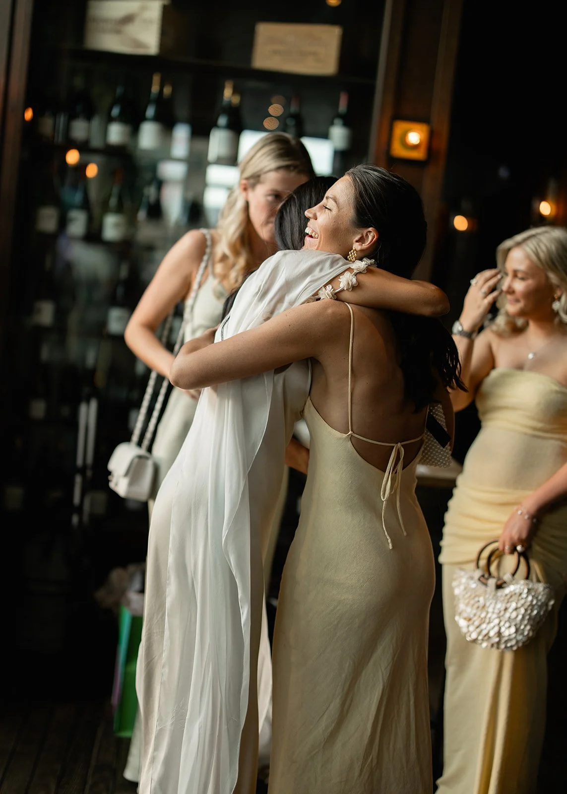 Two women hugging at a social gathering, one wearing a white dress and the other in a cream-colored dress, with other women in the background.