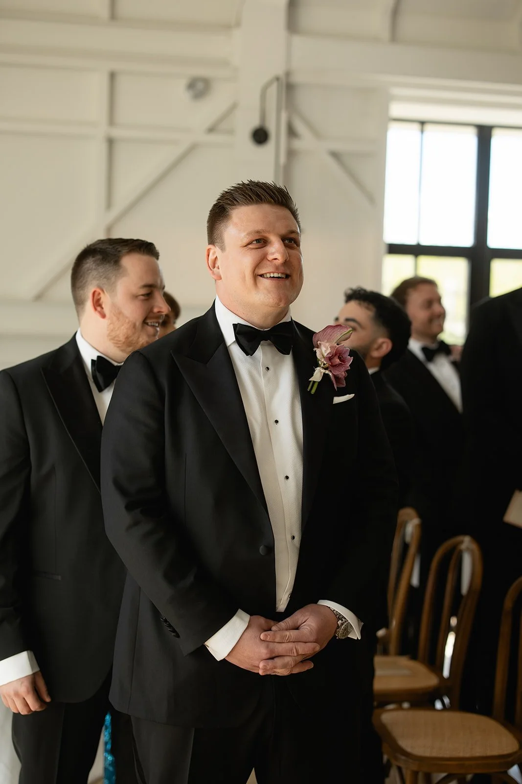 A groom in a black tuxedo and bow tie standing with his hands clasped, smiling during a formal ceremony with groomsmen dressed similarly in tuxedos, indoors near large windows.