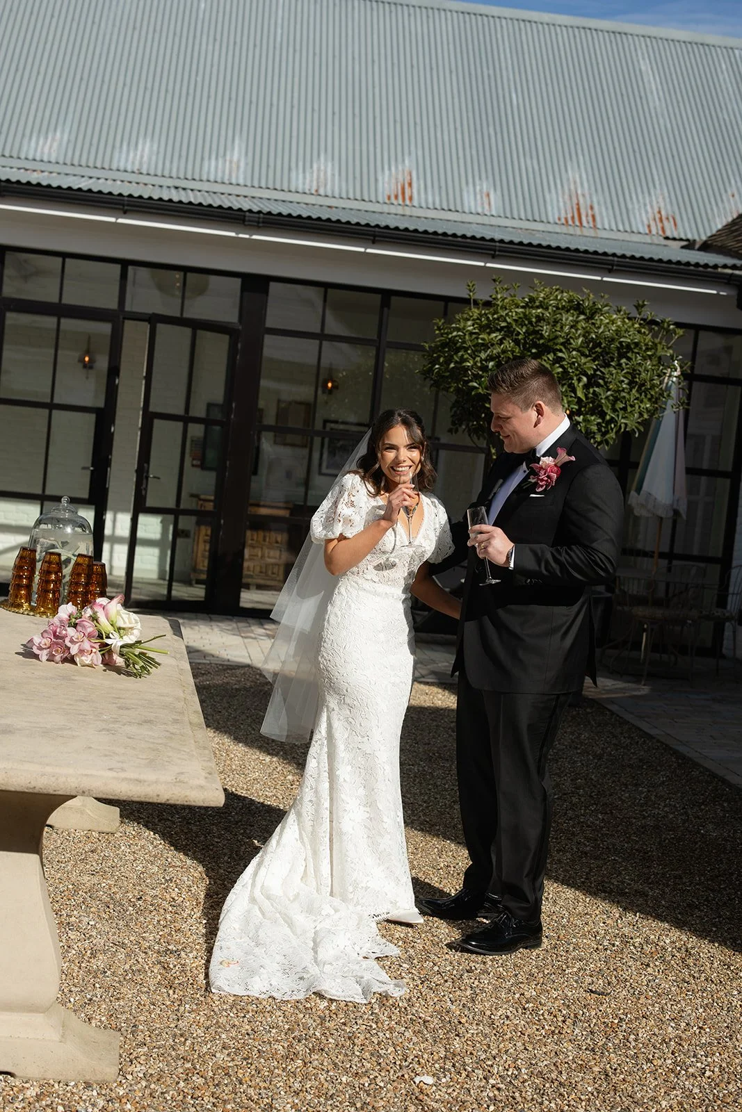 Bride and groom in wedding attire celebrating outdoors, toasting with drinks, smiling, with a table of beverages and flowers nearby.