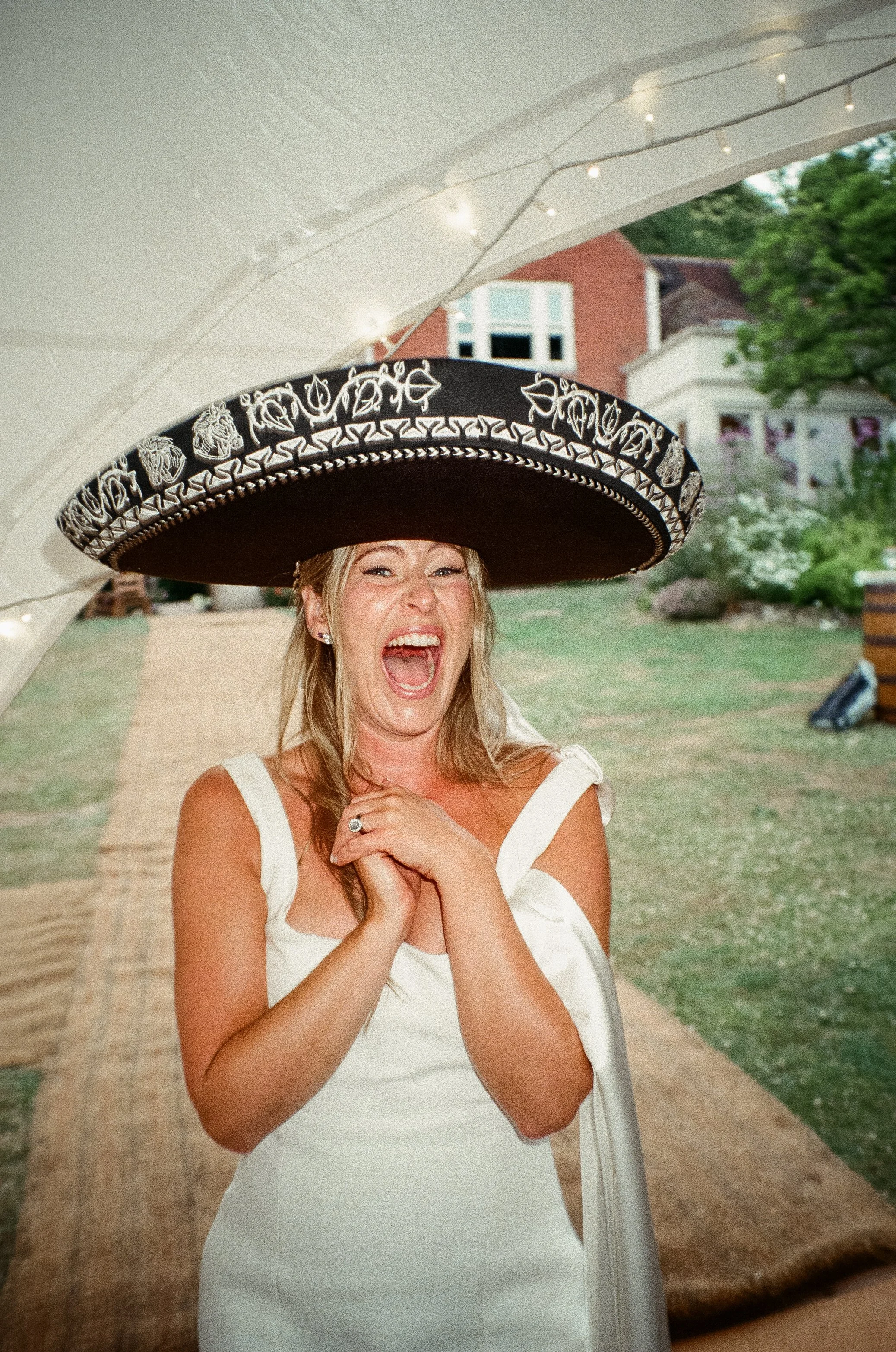 A woman in a white dress is wearing a large black and white sombrero and smiling with her mouth open, underneath a white canopy with string lights.