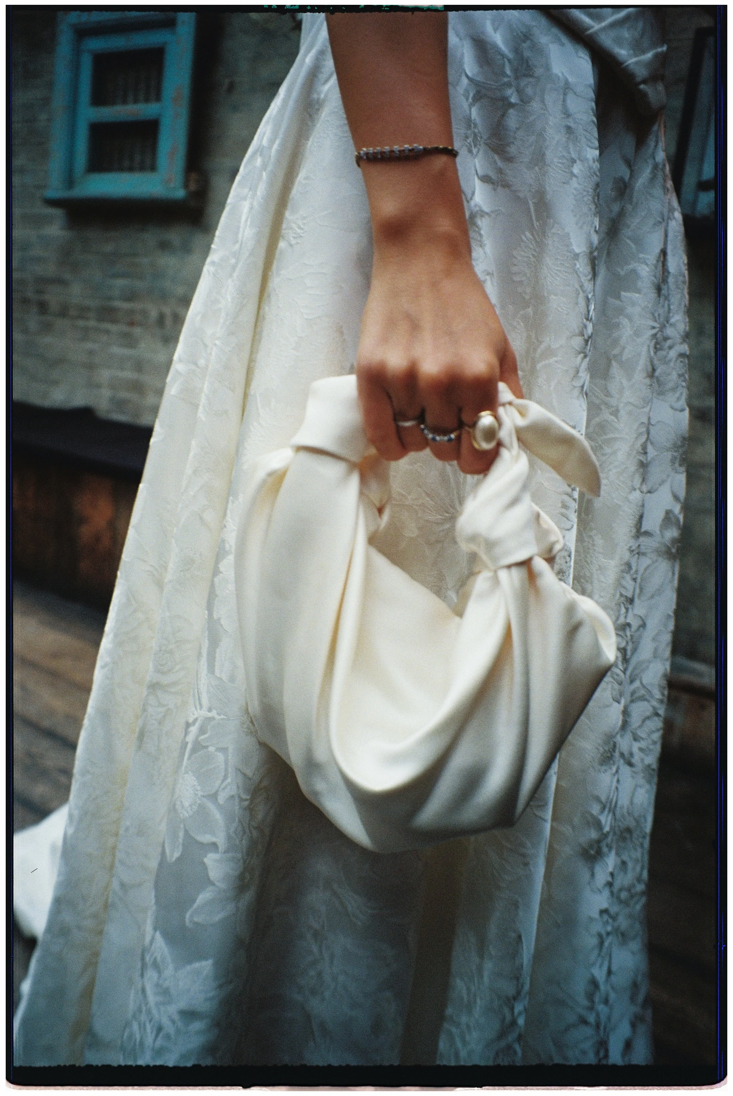 Close-up of a woman's hand holding a cream-colored satin clutch purse, she is wearing rings and a bracelet, and a cream lace dress.