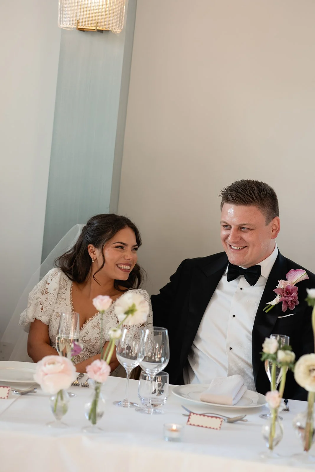 A bride and groom sitting at a decorated wedding table, smiling and looking at each other, with flowers and glassware in front of them.