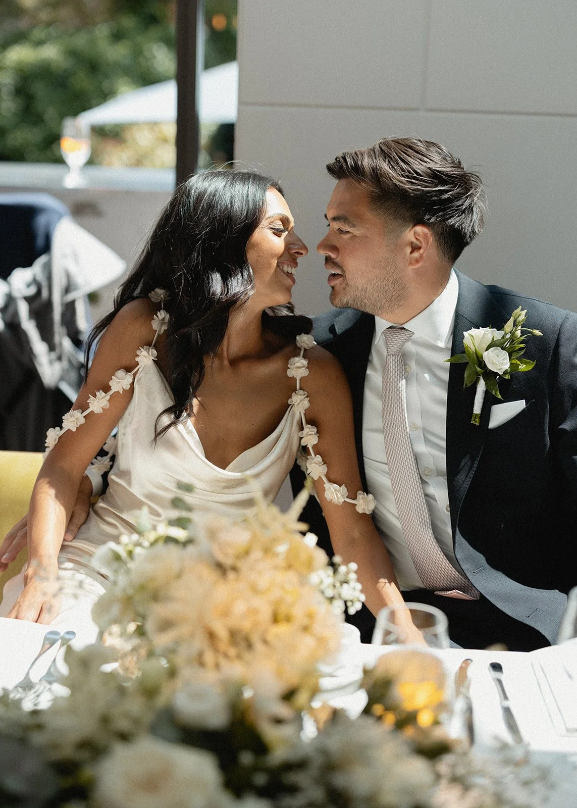 A bride and groom sitting close together at their wedding reception, smiling and leaning towards each other with foreheads touching.