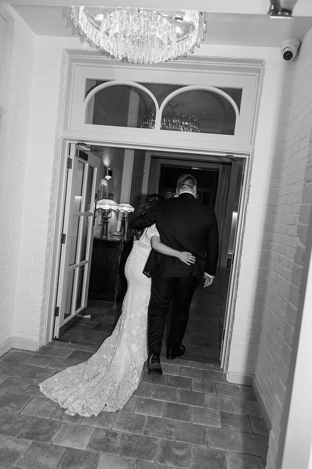 A bride and groom walking through a doorway at their wedding reception, with the bride in a lace wedding gown and the groom in a suit, embracing each other.