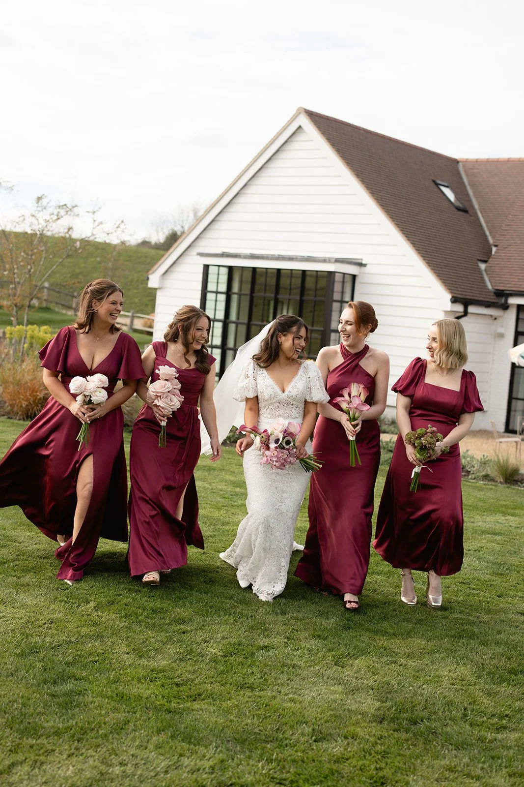 Group of women in burgundy bridesmaids' dresses and a bride in a white lace gown, walking outdoors on grass, smiling and holding bouquets, with a white house in the background.
