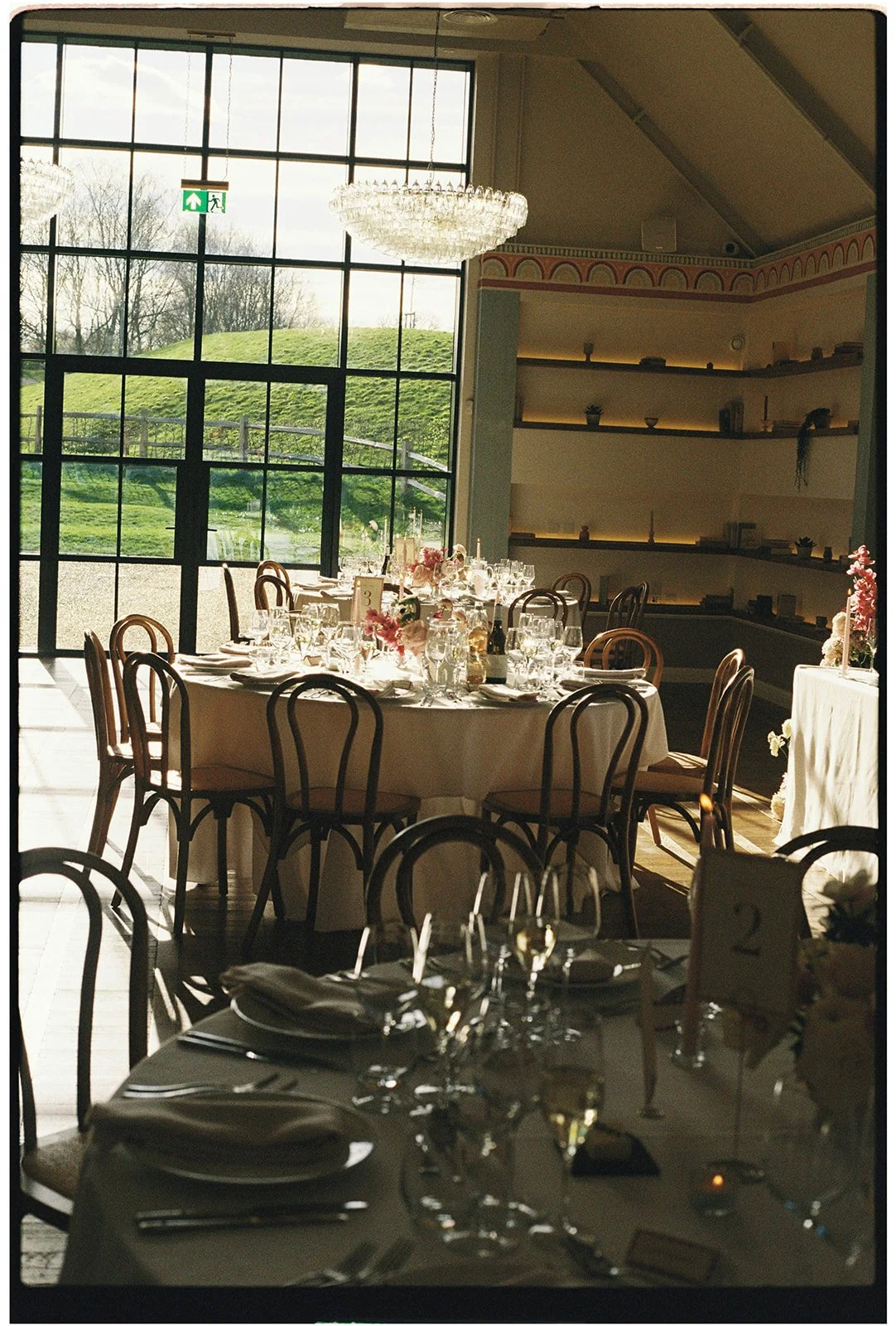 Elegant dining area with round tables covered in white tablecloths, set with glassware, napkins, and floral centerpieces. Large windows showcase a green outdoor landscape, and there is a chandelier hanging from the ceiling. The room is decorated with