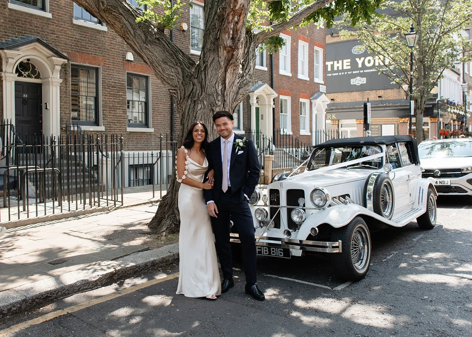 A newlywed couple standing beside a vintage white car parked on a city street, with brick buildings and trees in the background. The bride is wearing a white wedding dress and the groom a dark suit.