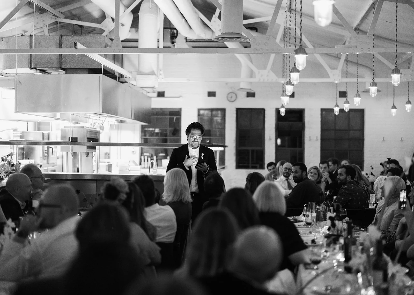 A black and white photo of a man standing in front of a seated audience in a modern restaurant or event space. The man is holding a microphone in one hand and a smartphone in the other, wearing glasses and a suit. The audience is engaged and looking towards him, while the room features an industrial design with high ceilings, hanging lights, and large windows.