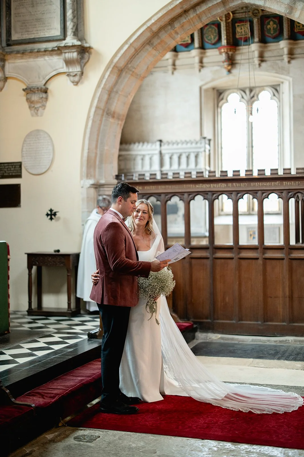 A bride and groom standing inside a church during their wedding ceremony, reading vows, with a priest in the background.