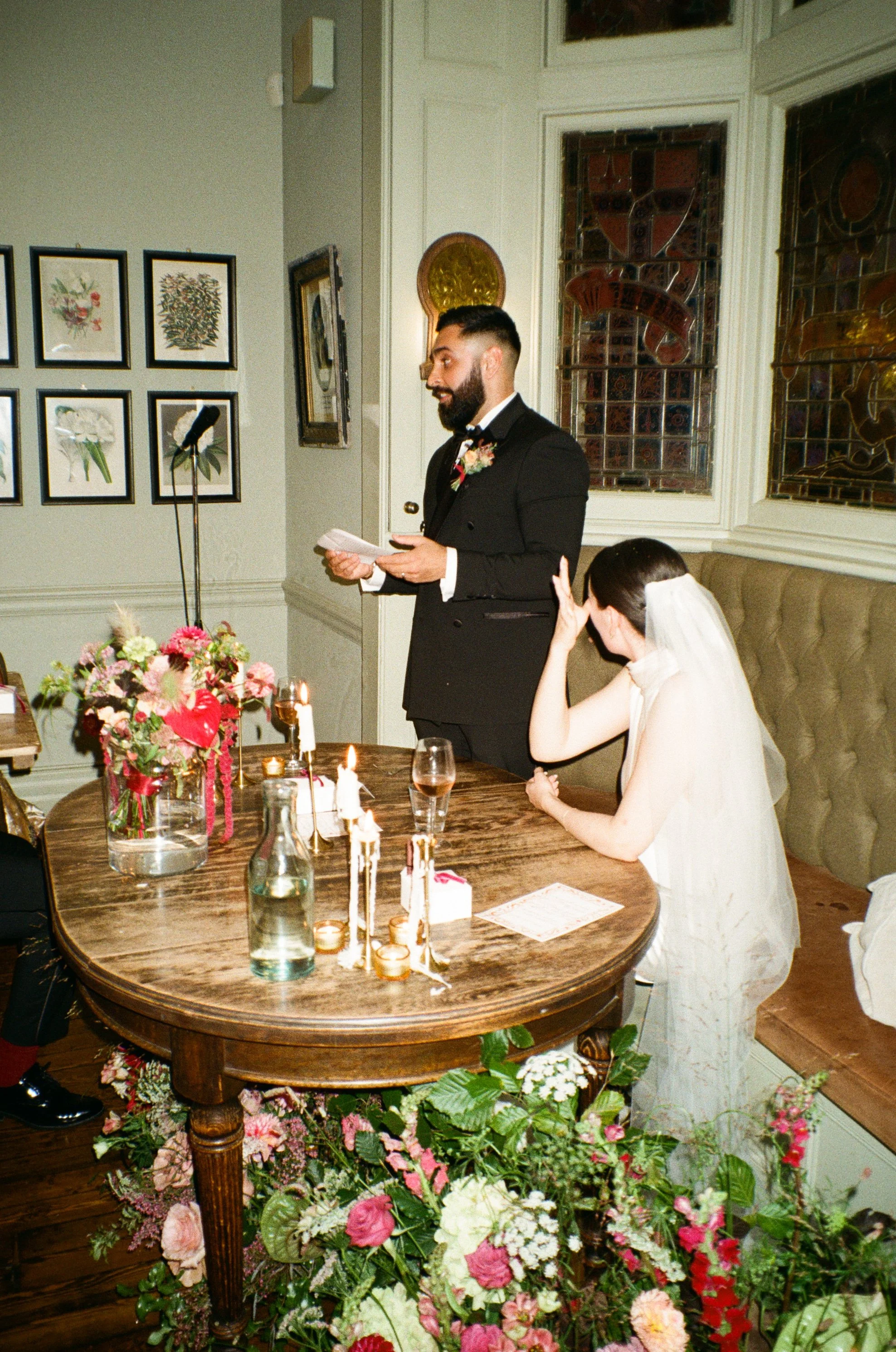 A bride and groom at their wedding ceremony; the groom is reading vows while the bride is seated, covering her face with her hand. The table is decorated with flowers, candles, and a flower arrangement, set in a room with art on the walls and stained