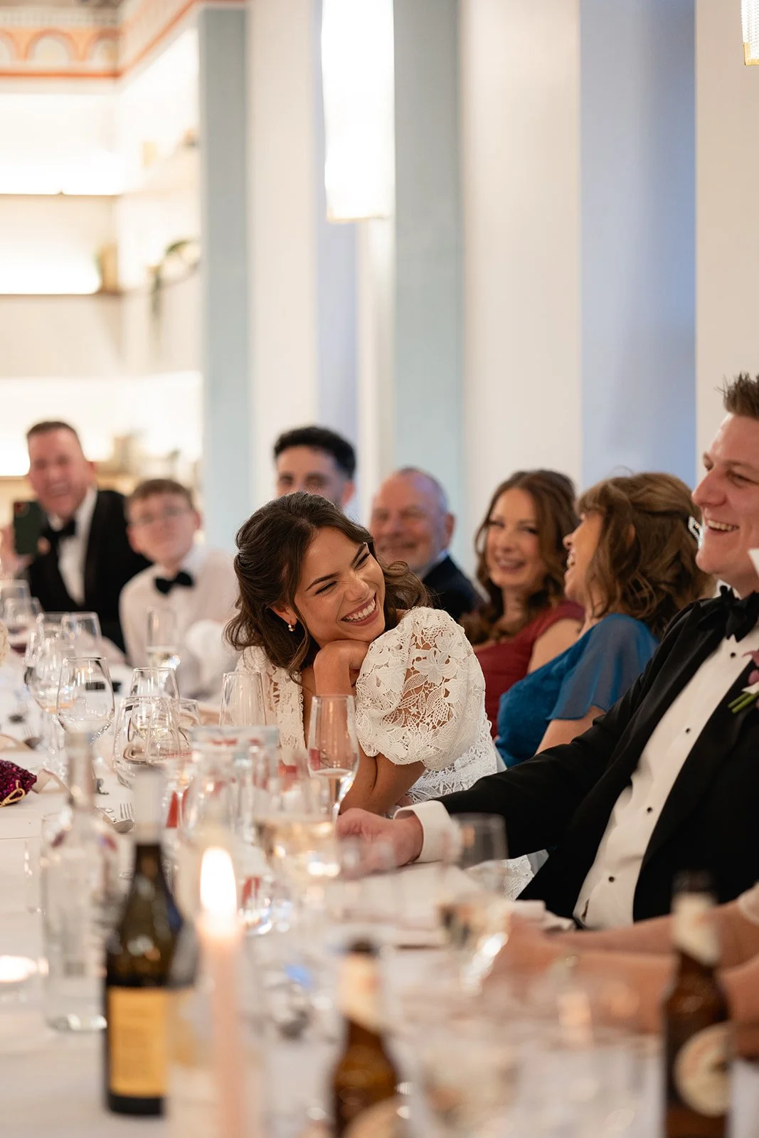 A woman in a white lace dress is laughing and leaning on a man in a tuxedo, at a formal dinner with other guests who are also smiling and talking.