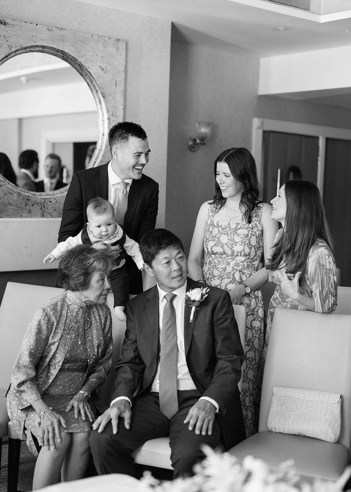 A group of people at a formal gathering, including a man in a suit, a woman in a floral dress, a woman in patterned dress, an elderly woman, a middle-aged man, and a baby, engaged in conversation and smiling.