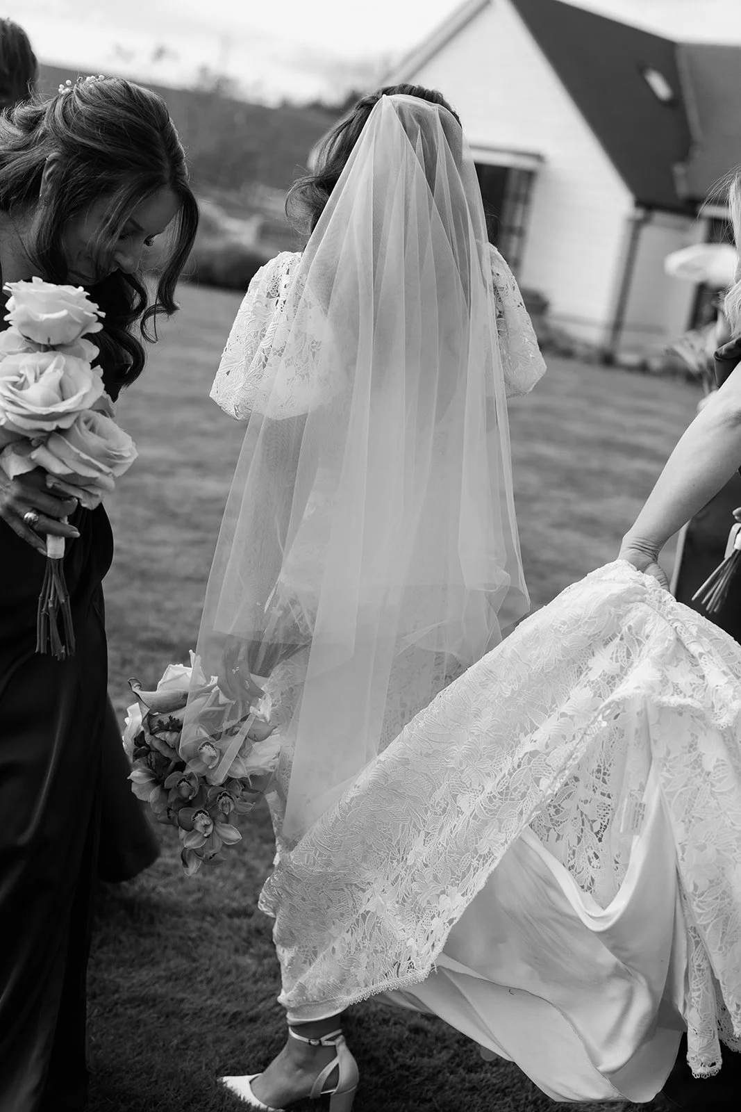 Black and white photo of a bride with a veil, surrounded by women, one holding a bouquet of roses, at an outdoor wedding.