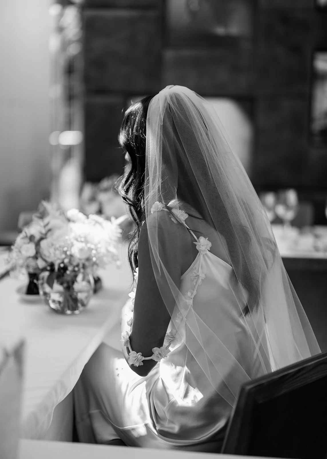 A bride with long dark hair wearing a wedding dress and veil, sitting at a table with a flower arrangement in a dimly lit room.