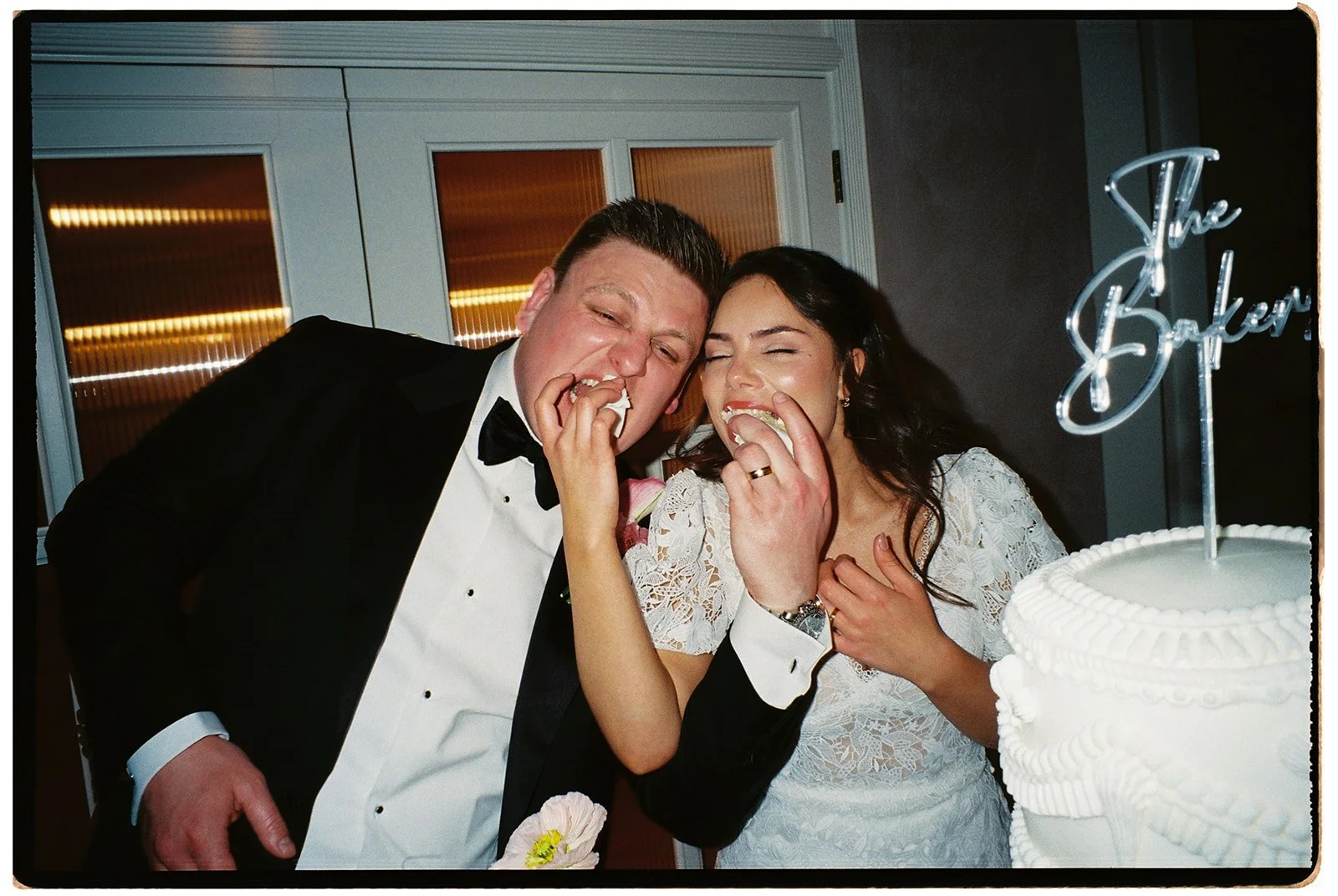A bride and groom in wedding attire eating cake together at their wedding reception, with a white wedding cake partially visible in the foreground.