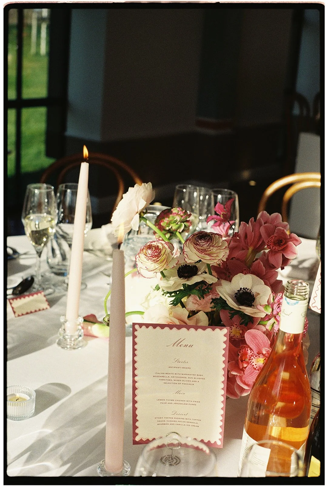 A decorated table with a floral centerpiece, a menu card, a lit pink candle, wine glasses, a bottle of rosé, and a small candle in a glass holder. The background shows chairs and a window with a view outside.