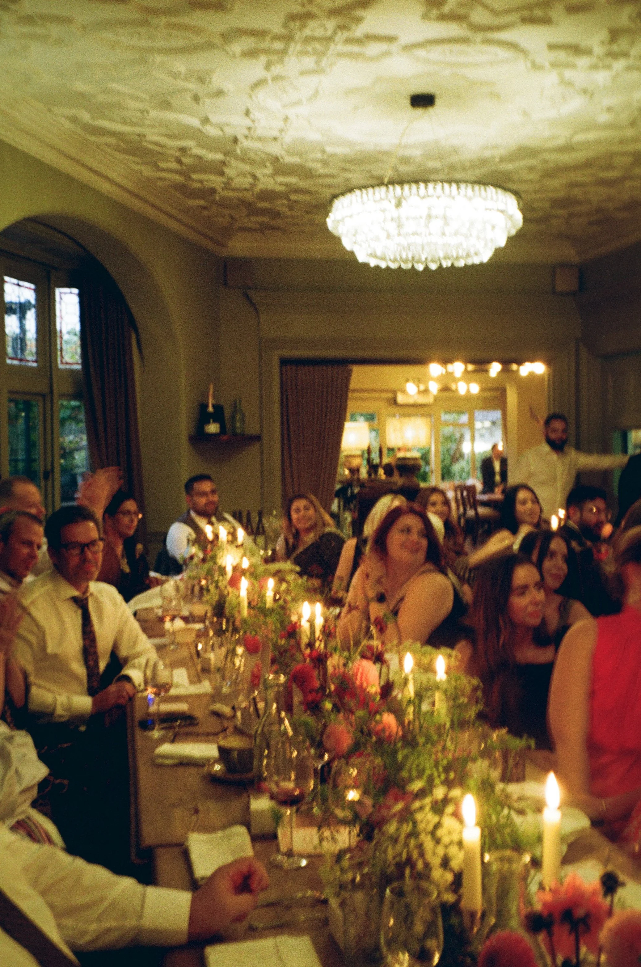 A group of people gathered around a long dining table decorated with flowers and candles at a formal event in an elegant room with a chandelier and large windows.