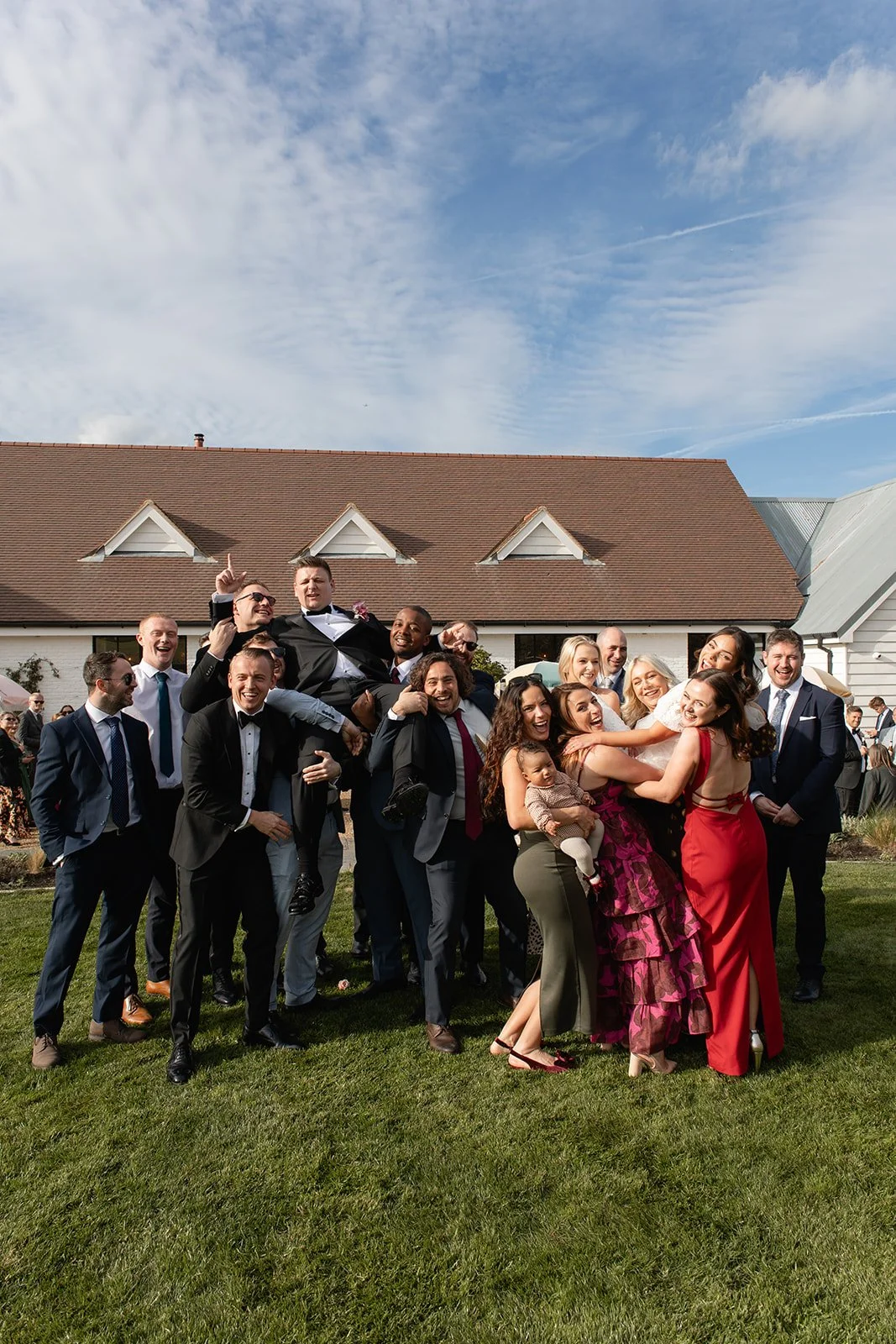 Group of people celebrating at a wedding, with some lifting the groom, outside on a grassy area near a building with a red-tiled roof.