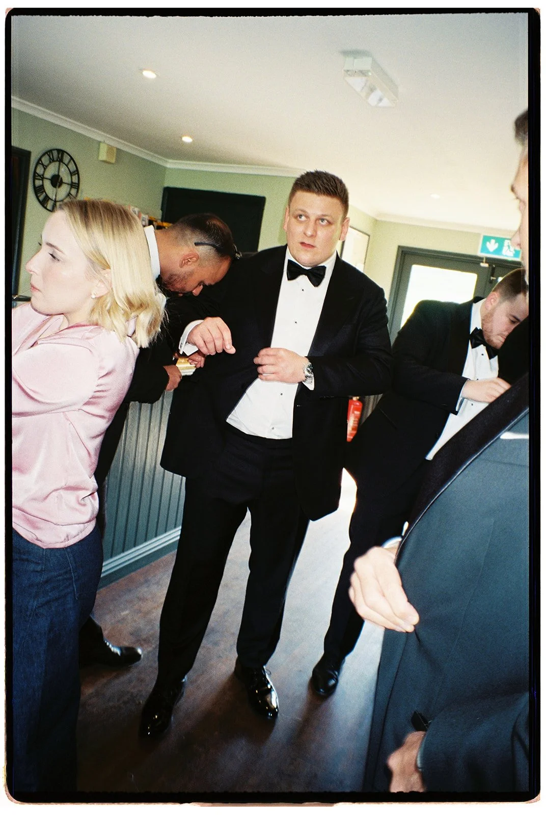 People dressed in formal attire, including tuxedos and a pink satin blazer, are gathered indoors, possibly preparing for an event or celebration.
