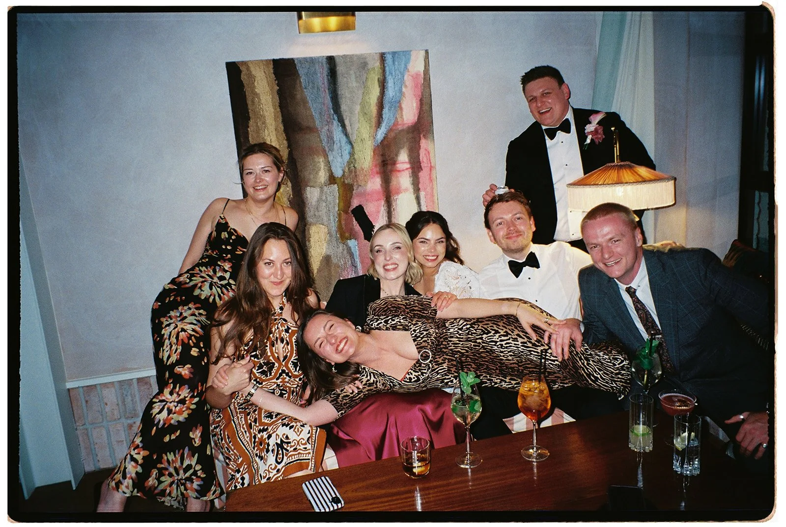 a 35mm analogue photo. Group of seven people celebrating at a formal gathering, some wearing tuxedos and dresses, with drinks on a table in front of them and an abstract painting in the background. The venue is Limekiln