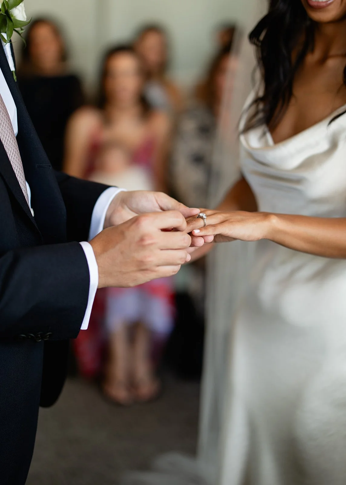 A person in a dark suit exchanges rings with a woman in a white dress during a wedding ceremony, with guests blurred in the background.