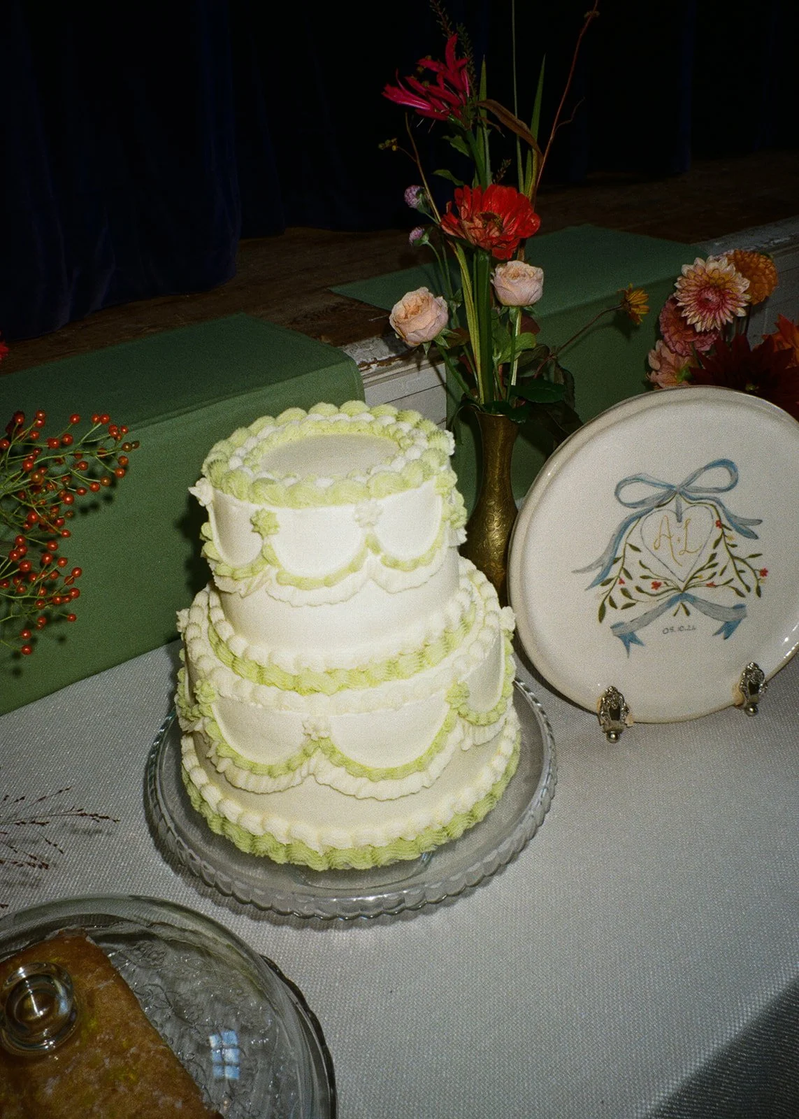 Three-tiered white wedding cake decorated with light green icing, placed on a glass plate on a table with a white tablecloth. Next to the cake is a white plate with a ribbon and floral design. Behind the cake are flowers in vases, including pink, red