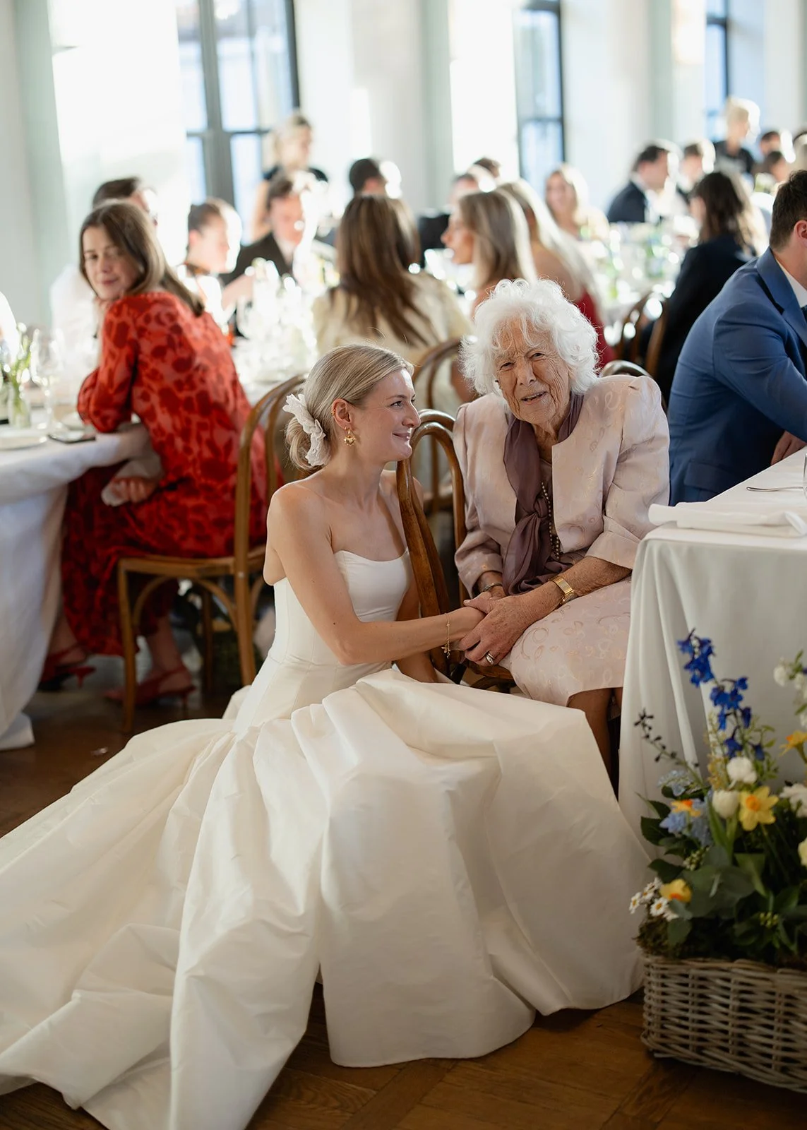 Bride in a white wedding dress sitting on the floor and holding hands with an elderly woman at a wedding reception.