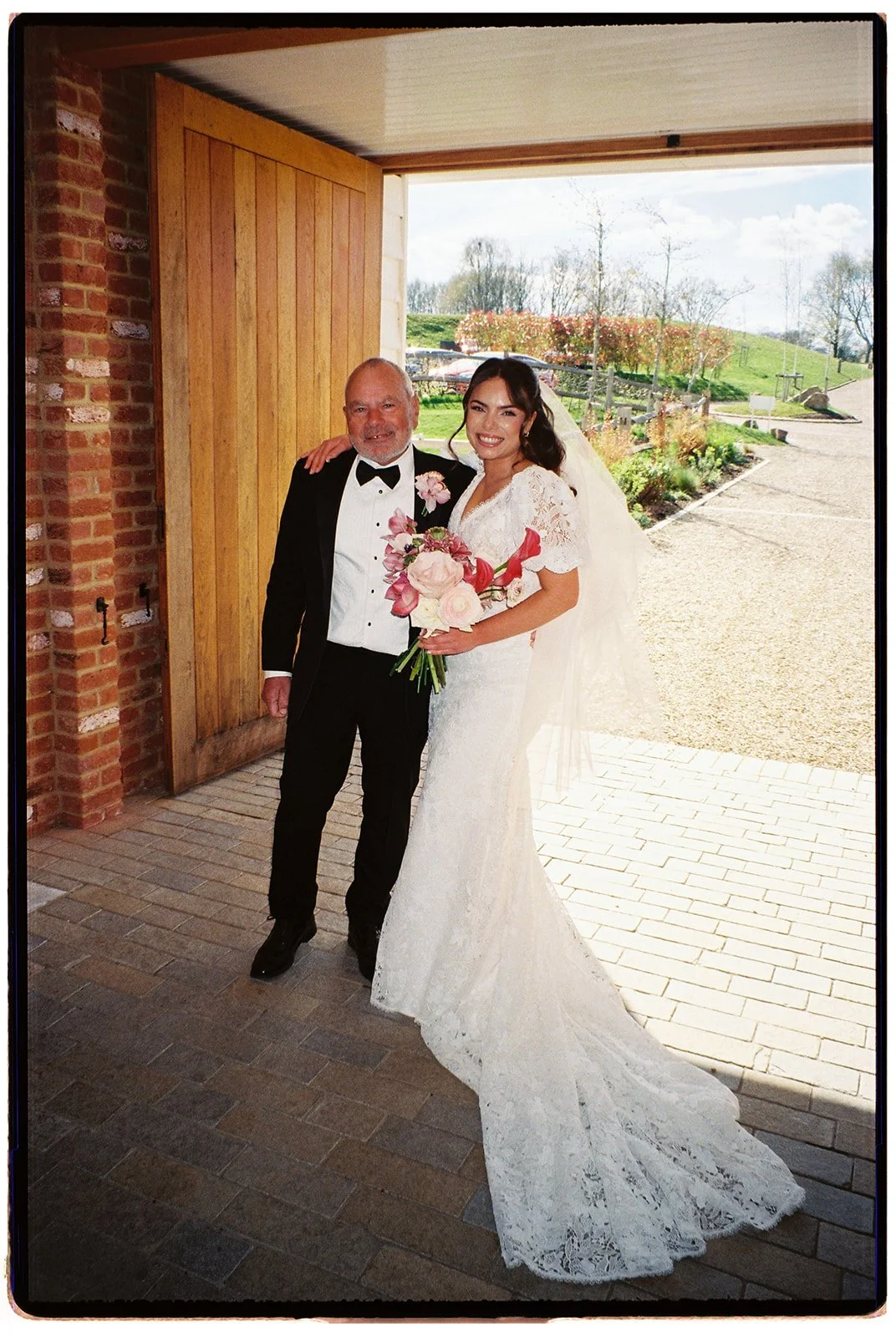 Bride in a white lace wedding dress holding a bouquet of pink and white flowers, standing next to an older man in a black tuxedo, inside a building with an open door showing a sunny outdoor landscape.
