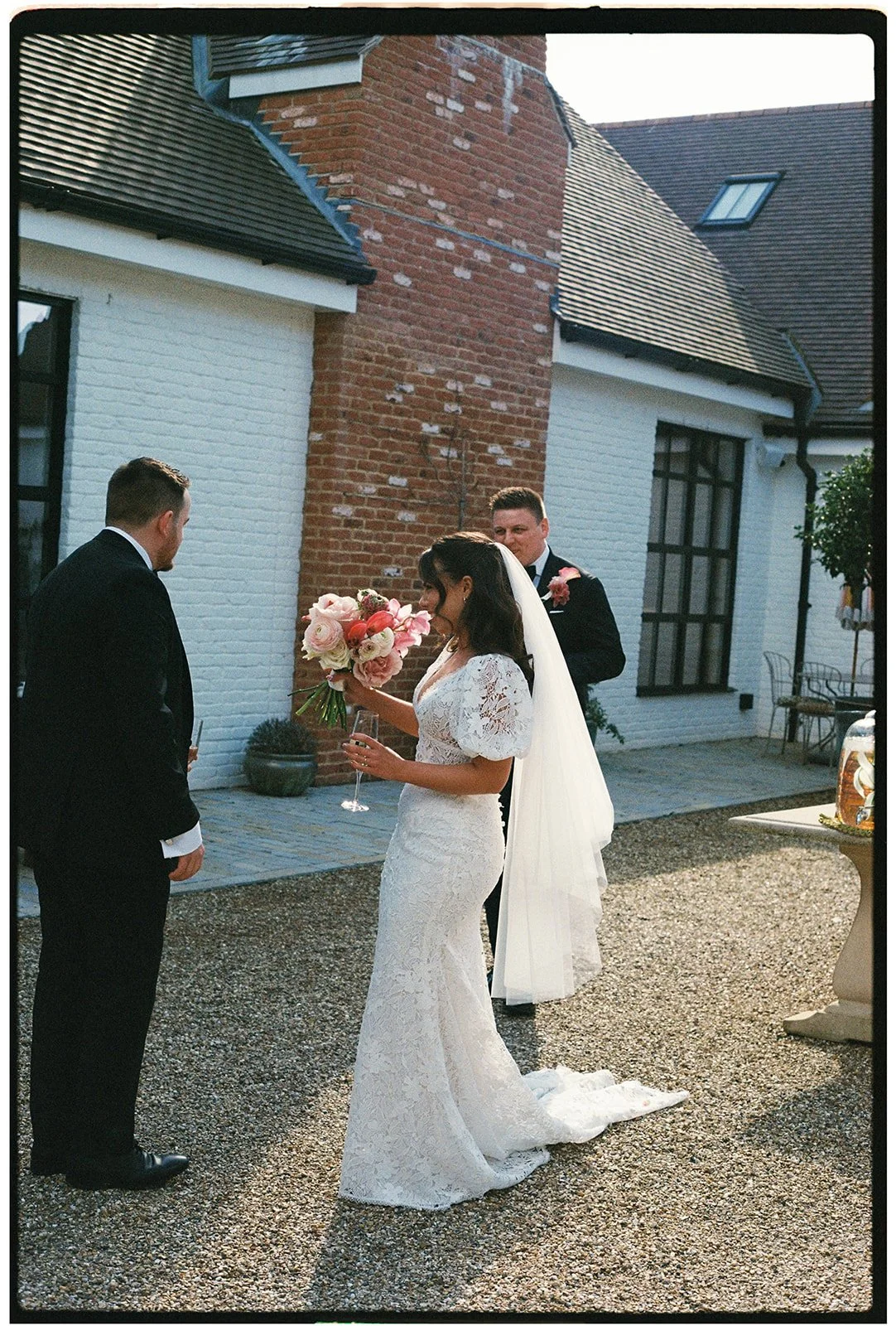Bride in a lace wedding dress holding a bouquet of pink and white flowers, standing outside with two men in tuxedos during a wedding ceremony.