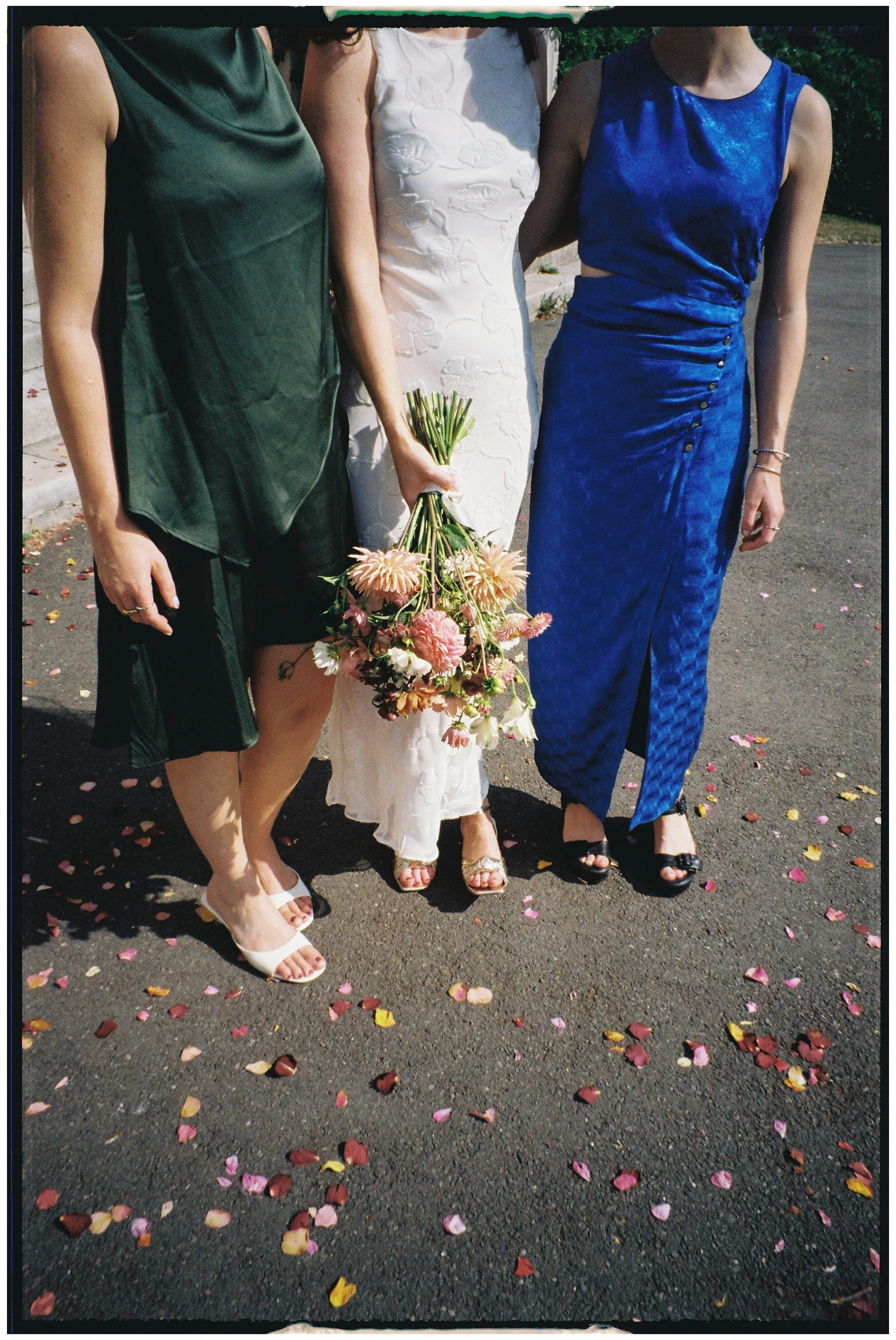 Three women standing outdoors on a paved area with colorful flower petals scattered around. The woman in the middle is holding a bouquet of pink and white flowers. The other two women wear elegant dresses: one in dark green, the other in vibrant blue