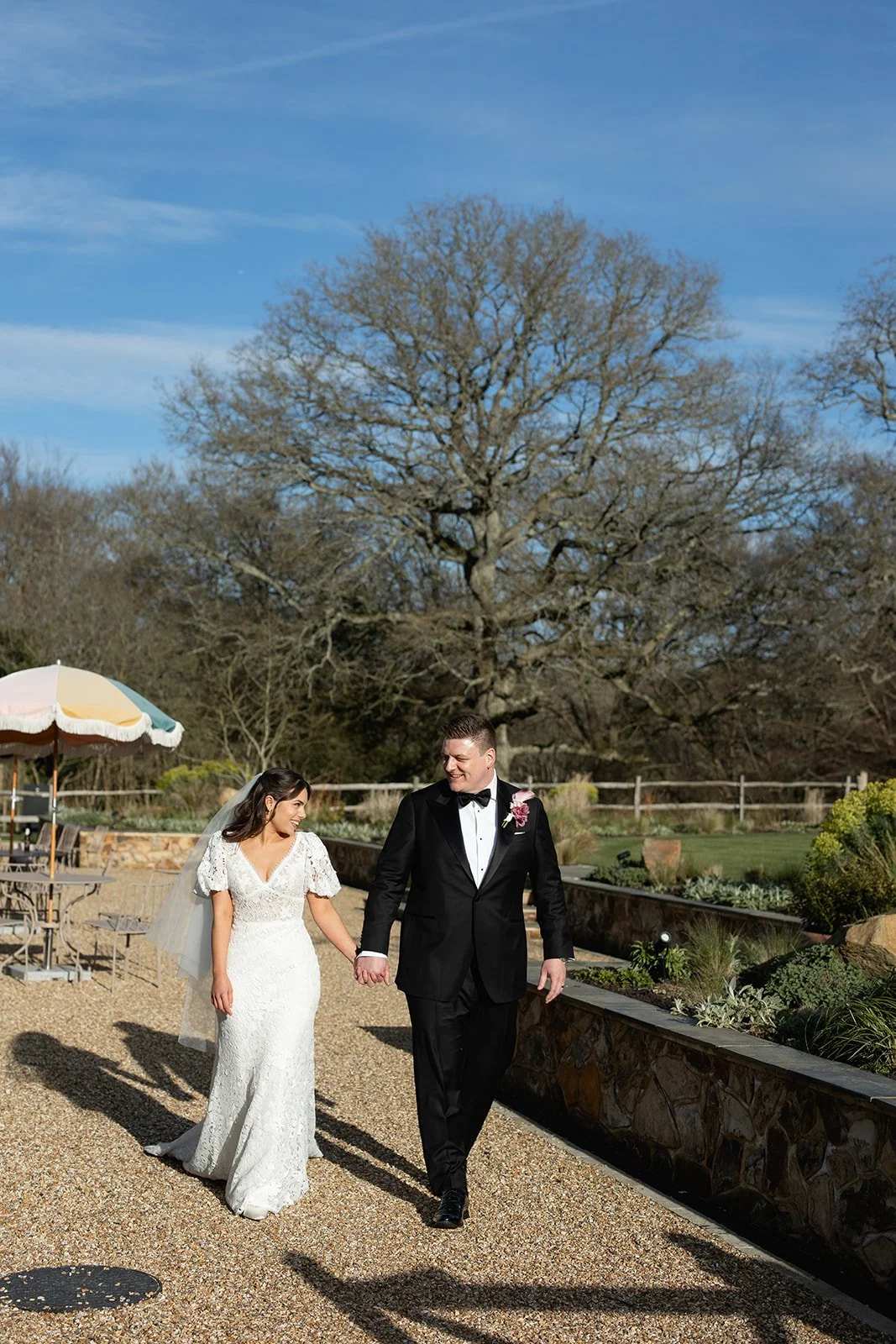 A bride and groom walking hand in hand outdoors on a sunny day, with a large tree and garden in the background.