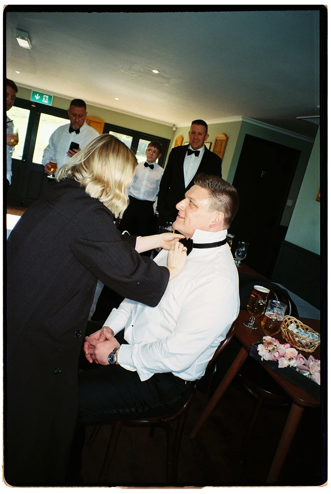 A woman helping a man with his tuxedo collar at a wedding reception. The man is sitting on a chair, smiling, wearing a white shirt and a black bow tie. Other guests in formal attire are in the background, with drinks on the table and pink flower peta