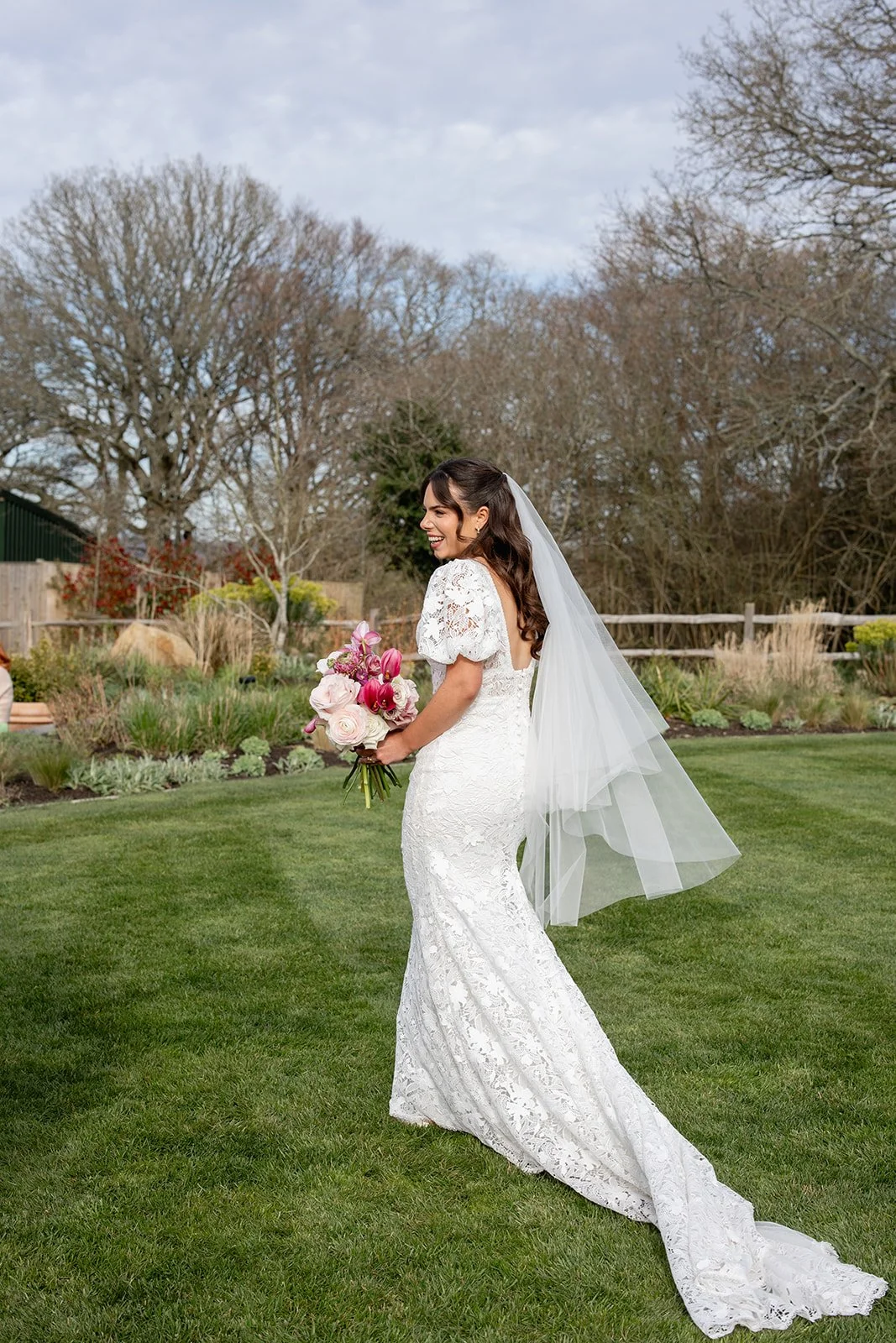 Bride in a white lace wedding dress with a long train and veil, holding a bouquet of pink and white flowers, standing on a green lawn with trees and shrubs in the background.