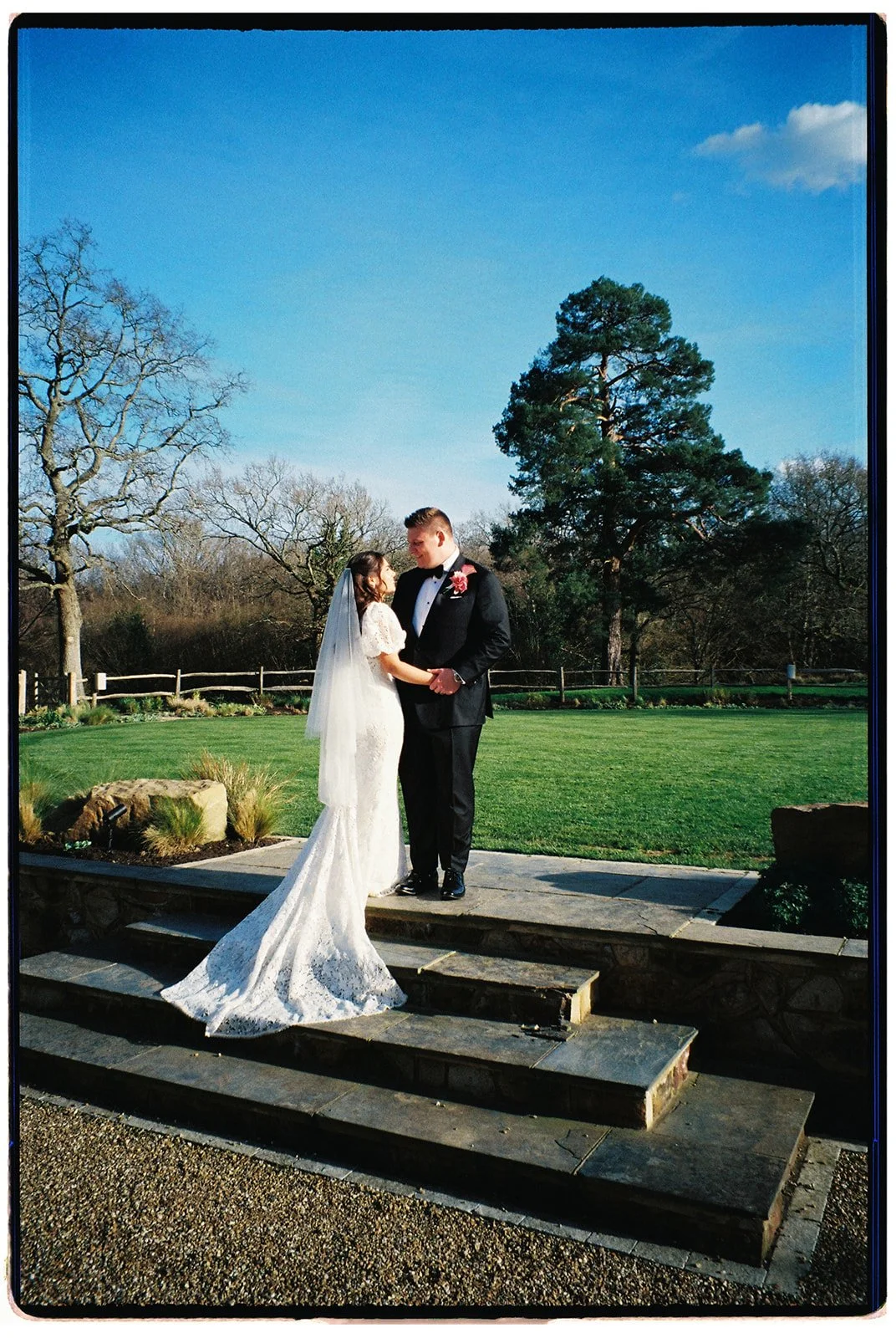 A bride and groom stand on outdoor stone steps, gazing at each other, with trees and a blue sky in the background, during their wedding photo shoot.