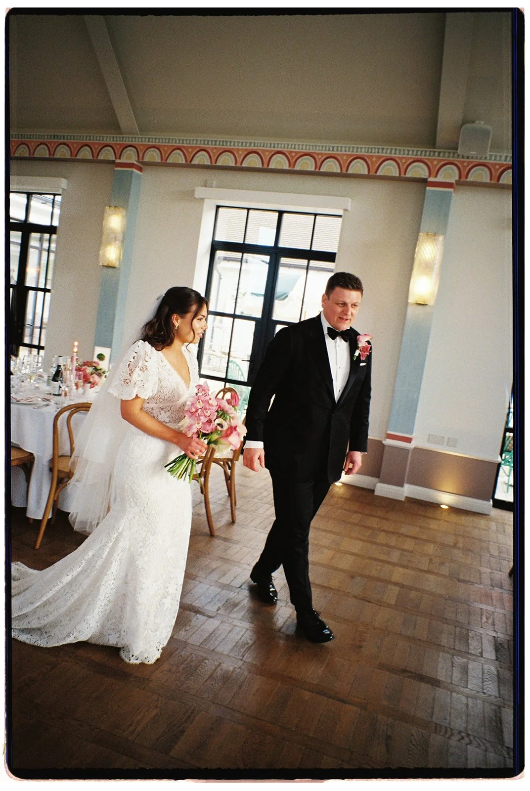 A bride and groom walking together inside a decorated wedding venue, with the bride holding a bouquet of pink flowers.