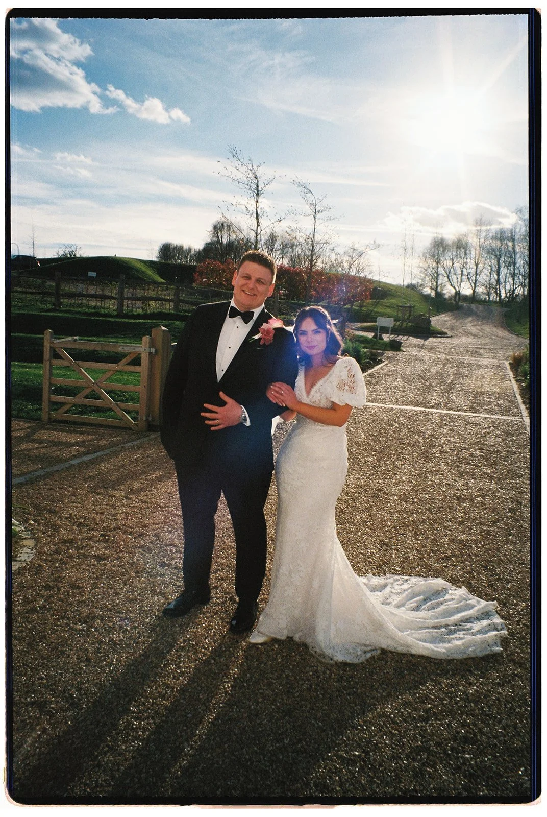 A bride and groom standing outdoors on a sunny day, dressed in wedding attire, with a scenic background of trees, a gravel path, and a partly cloudy sky.