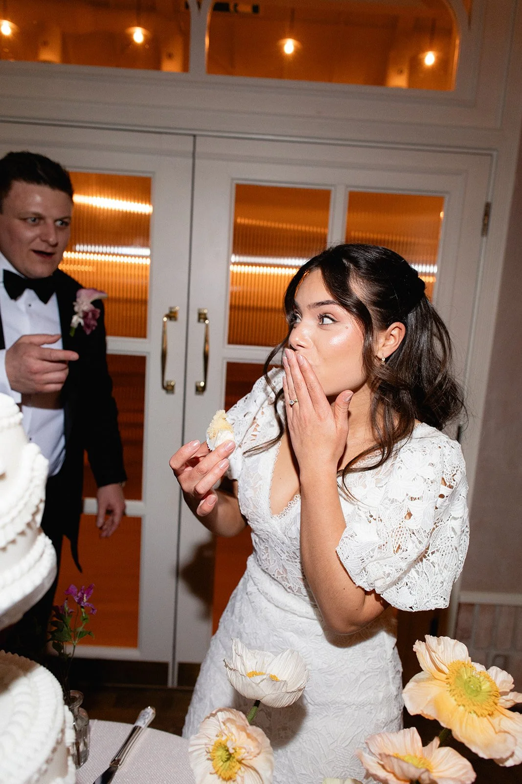 A bride in a white lace dress covering her mouth with one hand while holding cake in the other at her wedding reception, with a groom in a tuxedo pointing at her in the background. Flowers and a wedding cake are in the foreground.