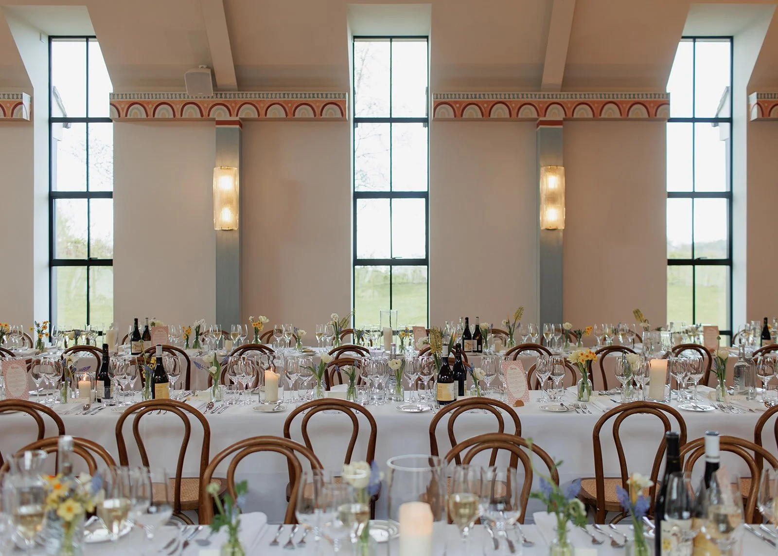 Elegant banquet hall set up for a wedding or celebration with long tables, floral centerpieces, candles, and wine bottles, large windows with natural light.