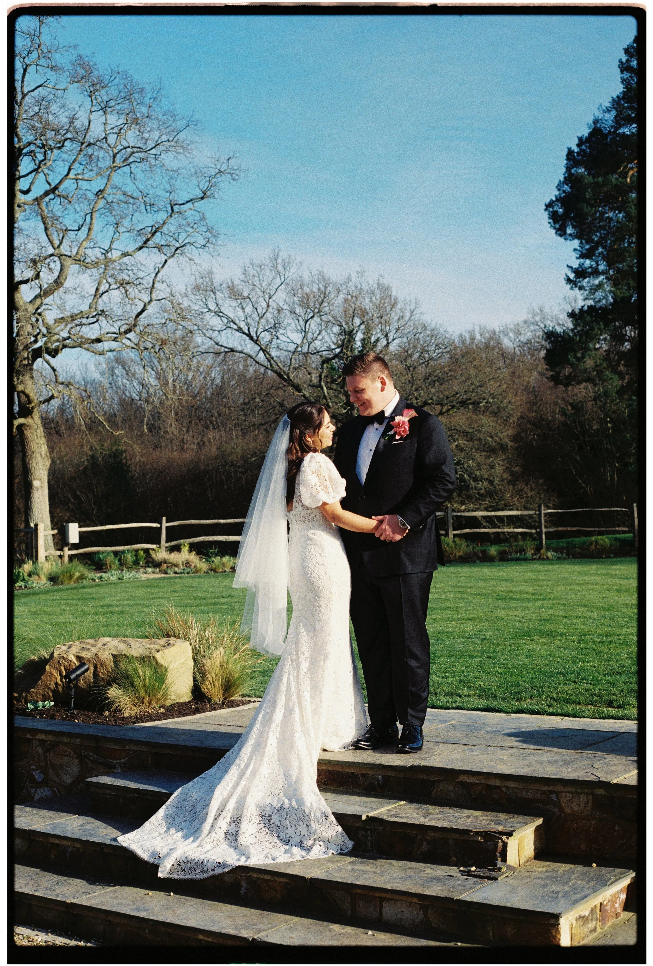 A bride and groom standing on steps outdoors, holding hands and sharing a moment during their wedding, with trees and a fence in the background on a sunny day.